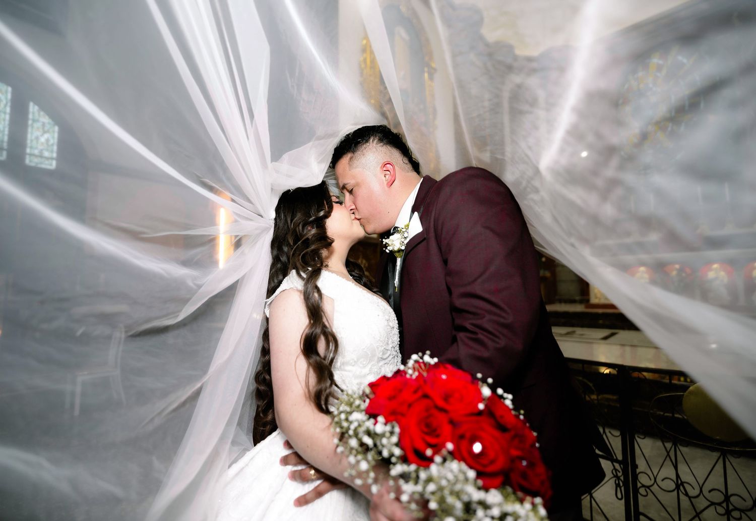 A romantic wedding moment captured under flowing white veils with a vibrant red rose bouquet.