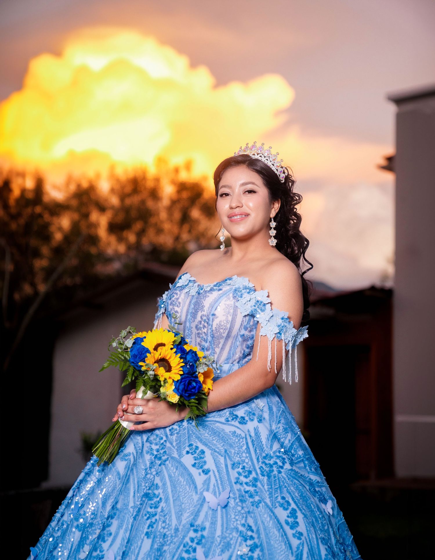 A person in a light blue quinceañera ball gown holds sunflowers while posing at sunset against a dramatic sky.