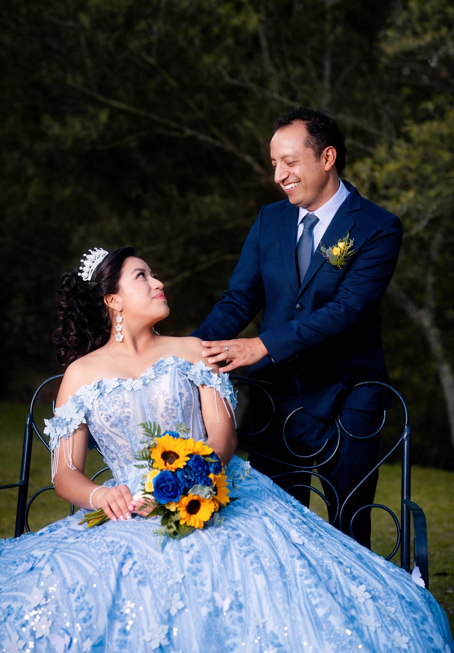 A couple in formal attire share a romantic moment, with sunflowers in the bouquet and a light blue off-shoulder ball gown.