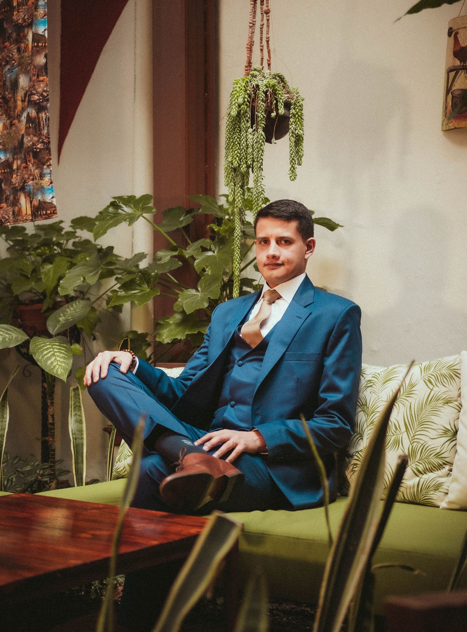 Figure in blue three-piece suit sits elegantly on green couch with hanging plants and decor in background.