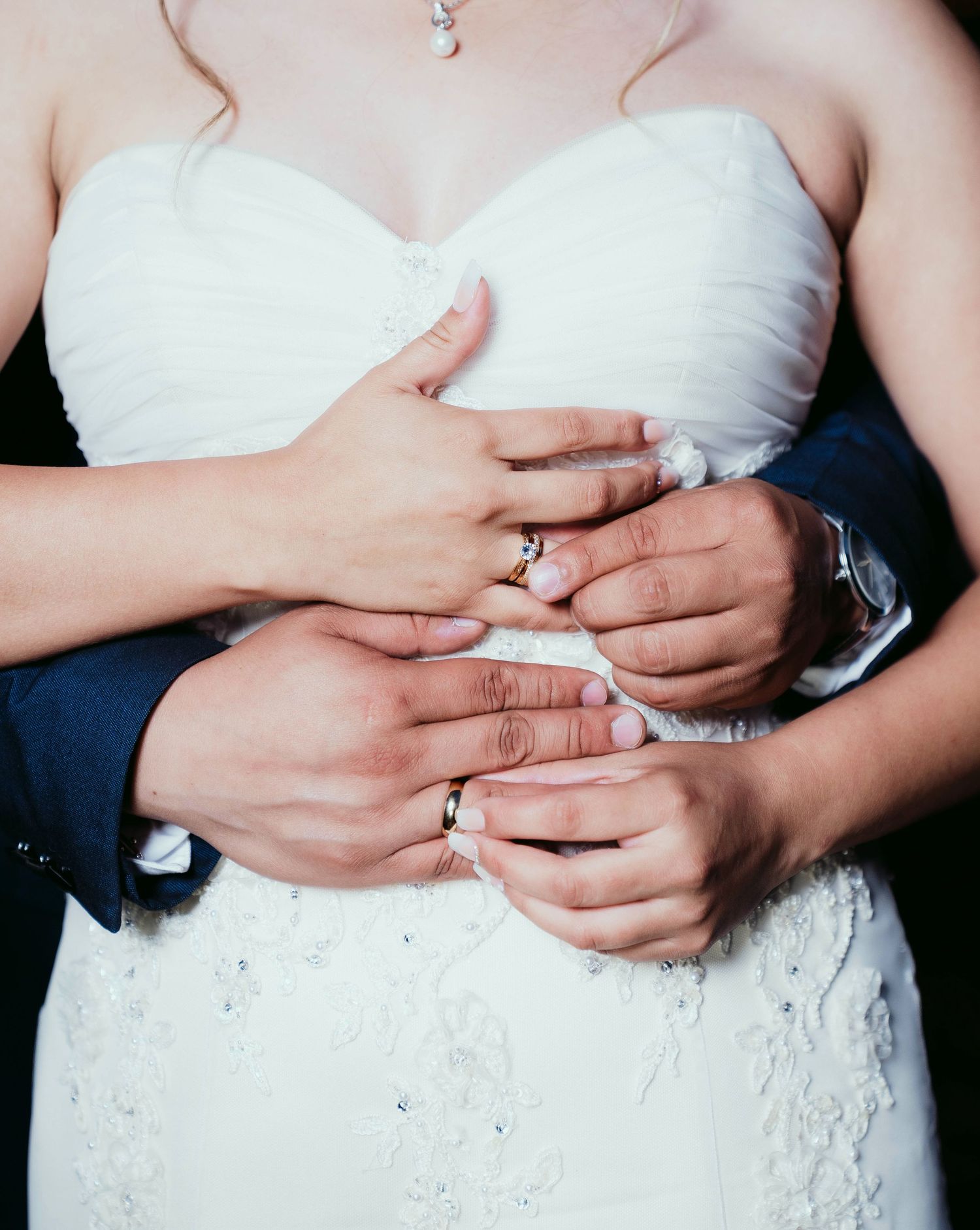 Close-up of hands clasped together showing wedding rings against a white bridal gown detail.