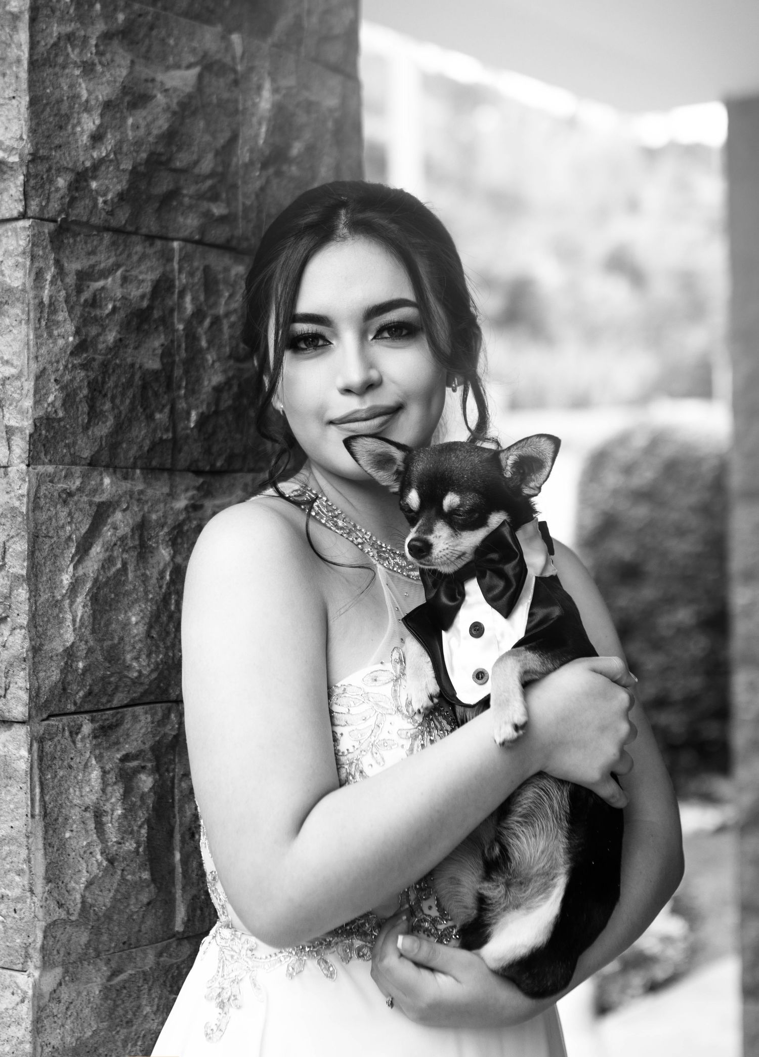 A bride in a white lace dress poses with her small dog wearing a tuxedo against a stone wall.