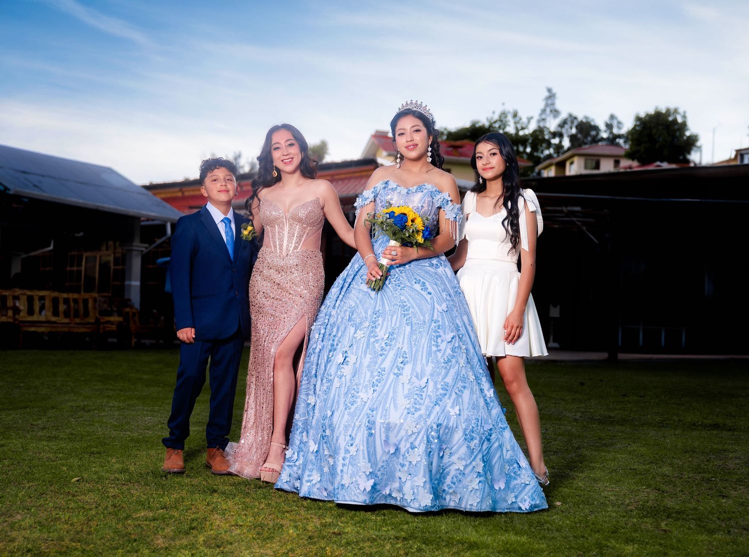 A group poses outdoors in formal attire including a blue ballgown and pink dress against a mountain backdrop.