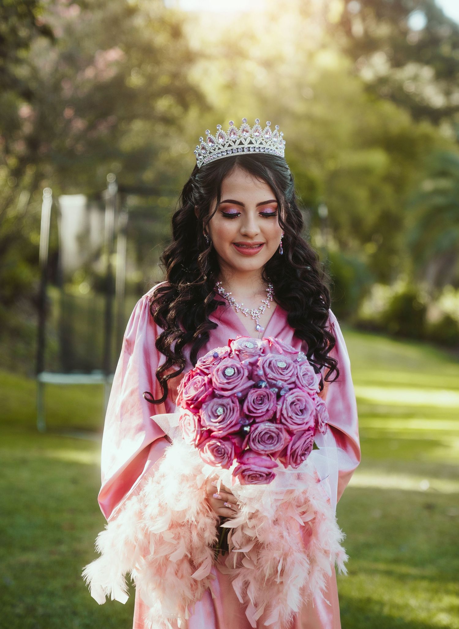 A person in a pink dress and tiara holds a bouquet of pink and purple roses in a sunlit outdoor garden setting.
