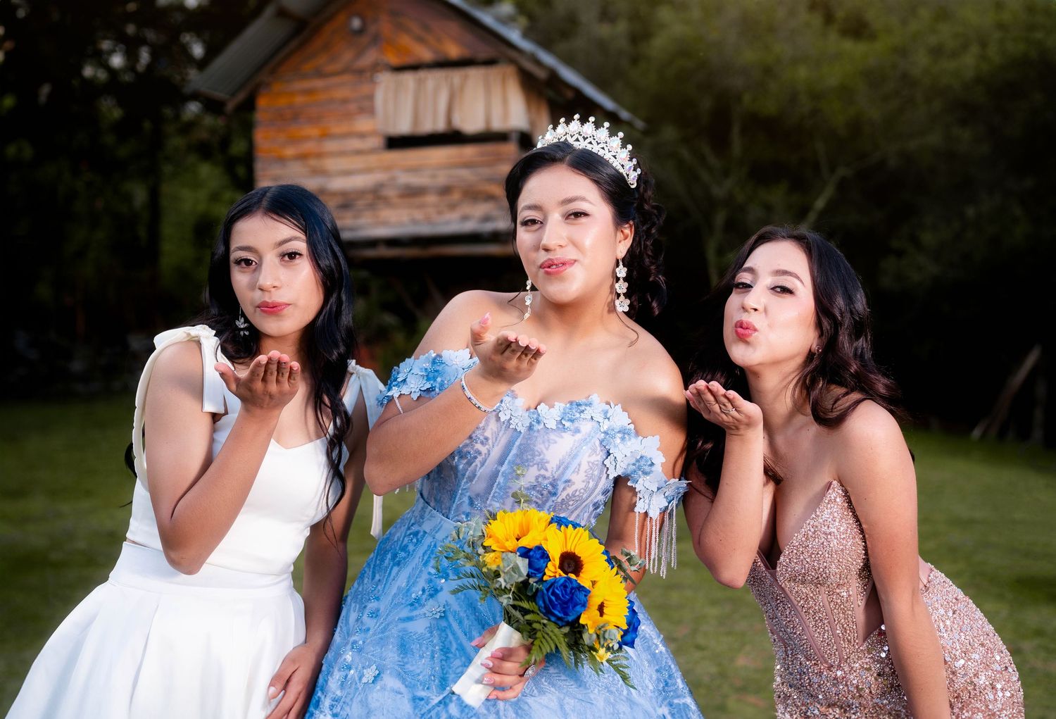 Three women in formal evening gowns pose together outdoors, with the center figure holding yellow sunflowers.