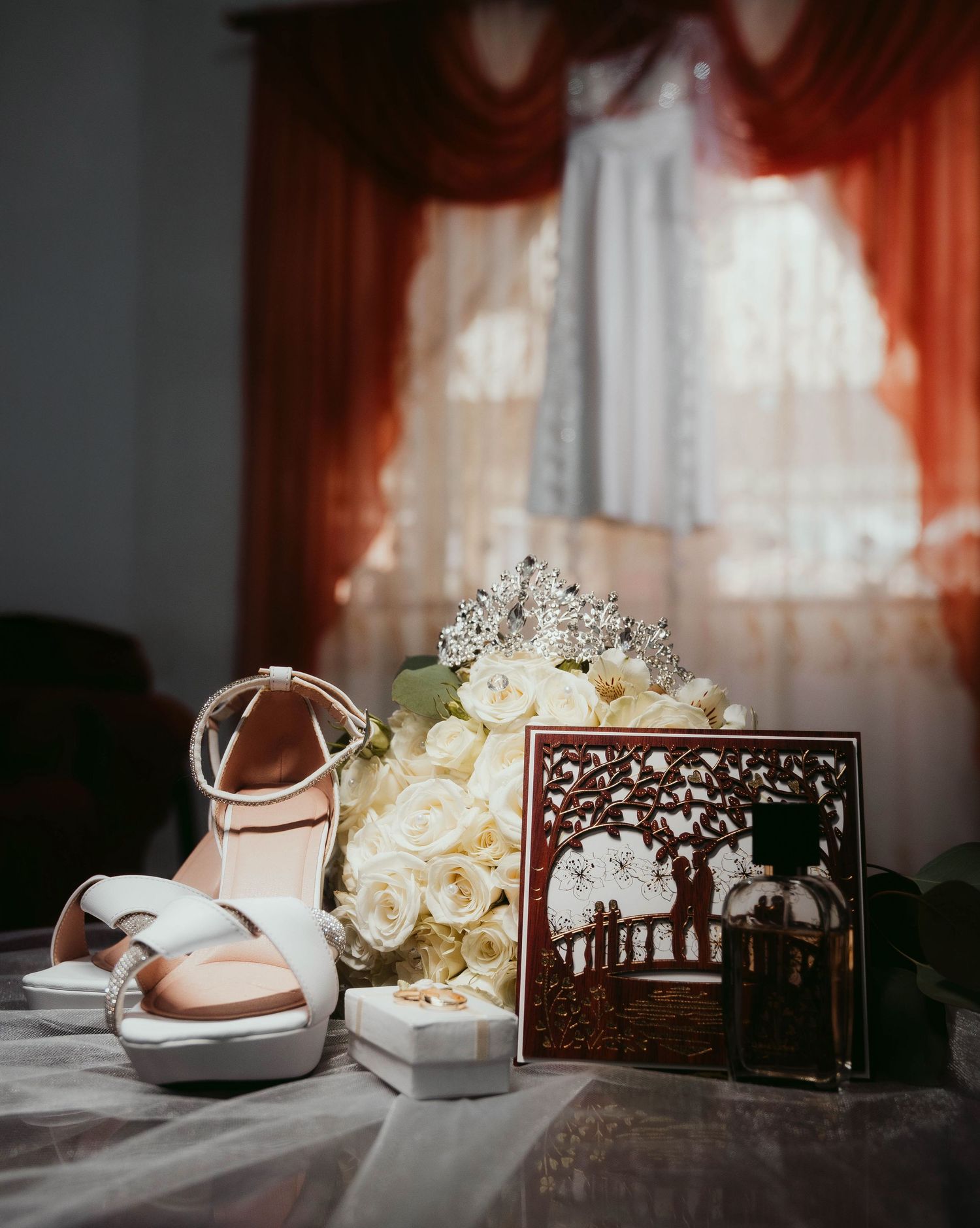 White bridal shoes, a floral bouquet, and a decorative laser-cut frame displayed on a table with red curtains in background.