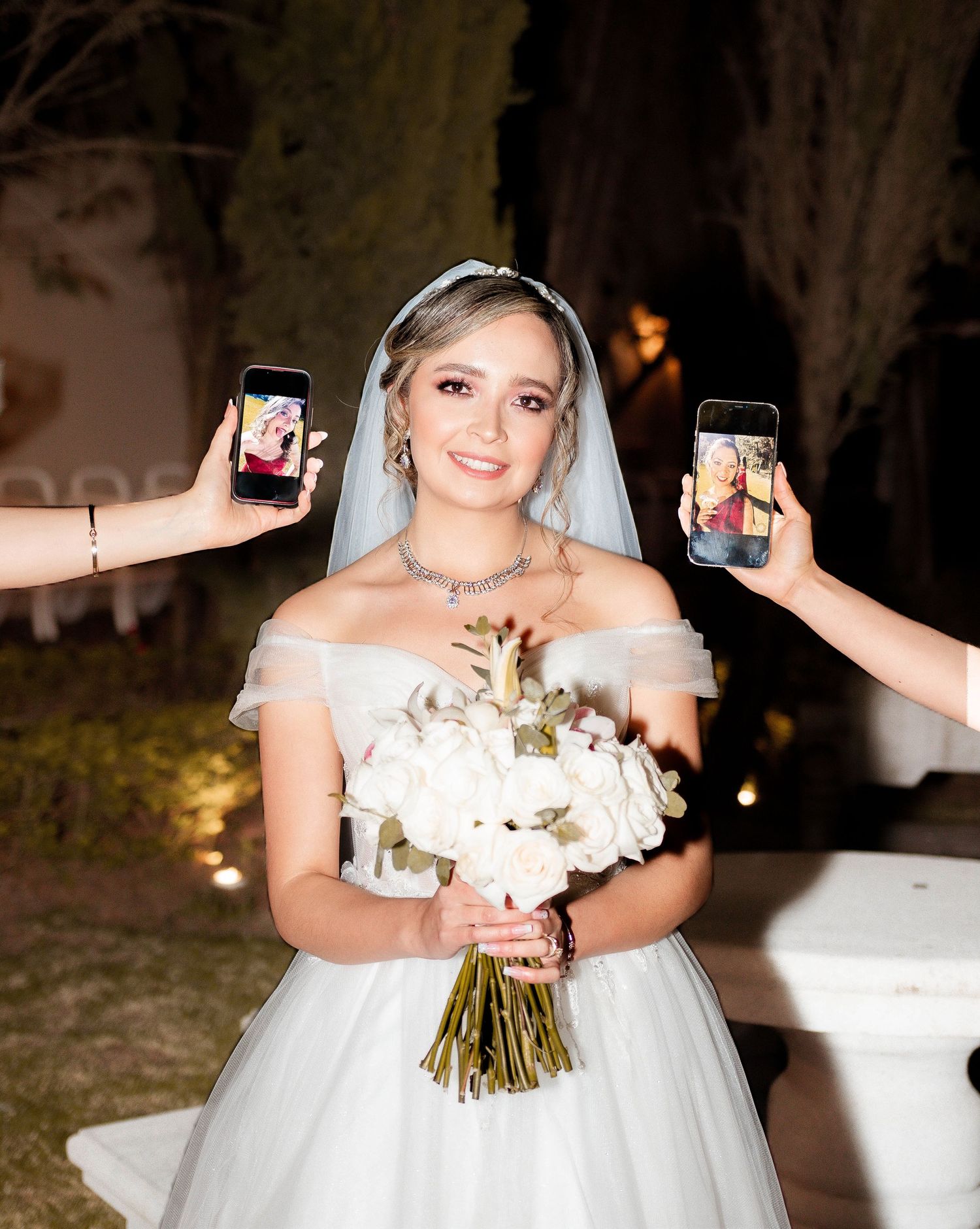 A bride in a white ballgown holds bouquet while guests take photos with phones.