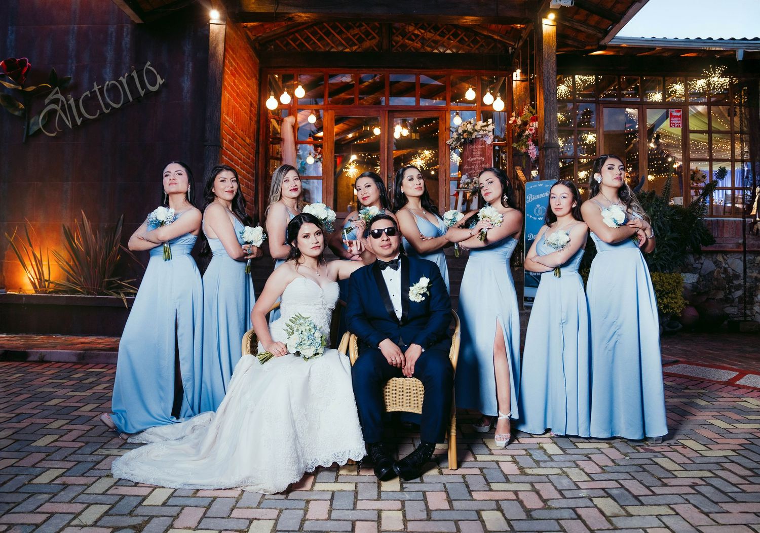 Wedding party poses together in evening lighting on brick patio outside reception venue with glowing lights.