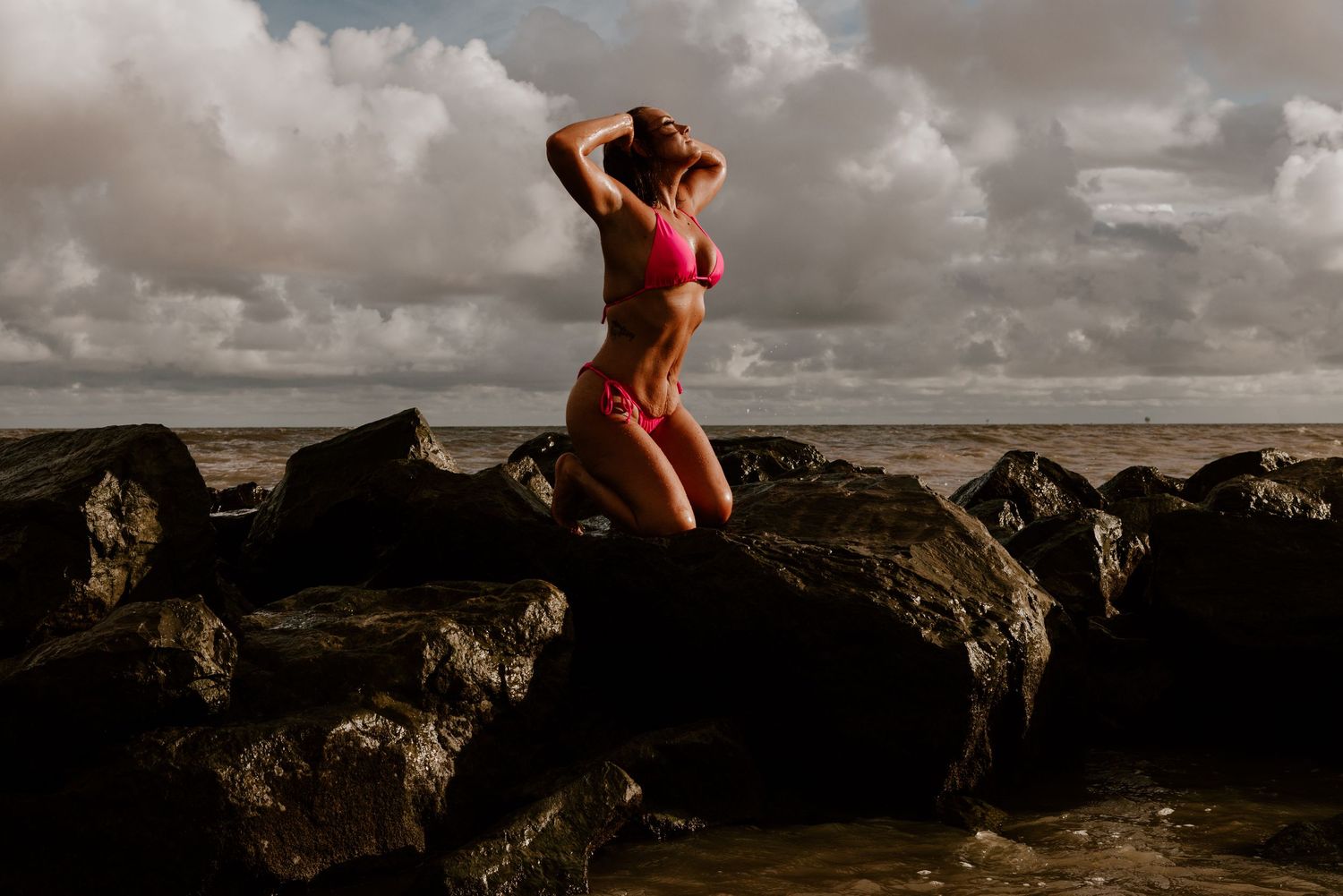 Figure in red swimwear poses on dramatic rocky coastline during moody sunset with cloudy skies and ocean backdrop.