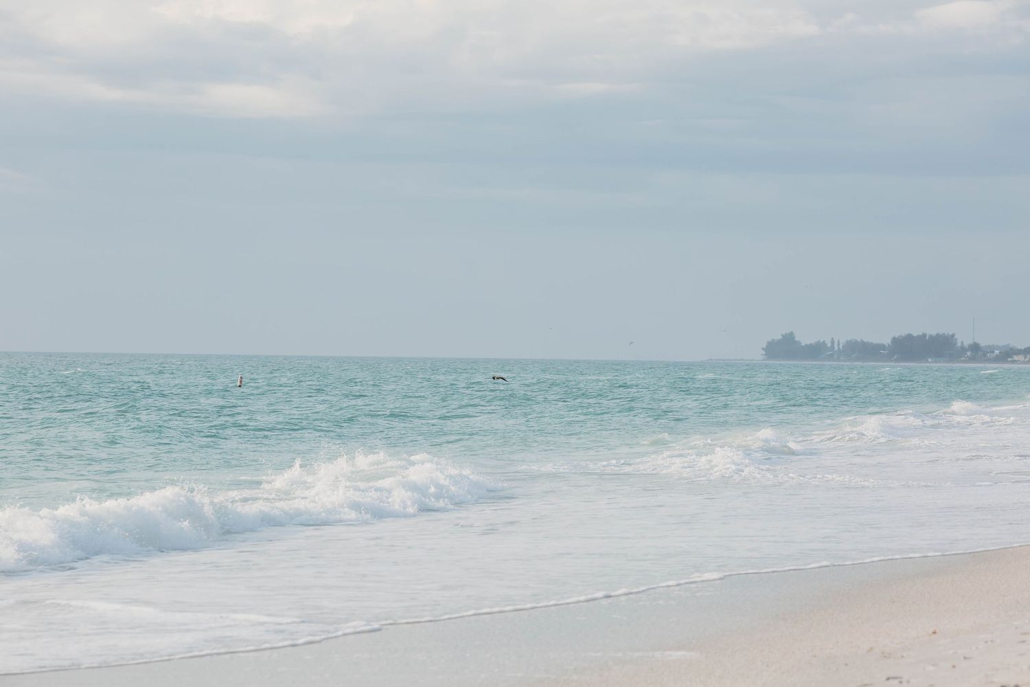 Serene turquoise ocean waves gently rolling onto a pristine white sand beach with distant buildings on the horizon.