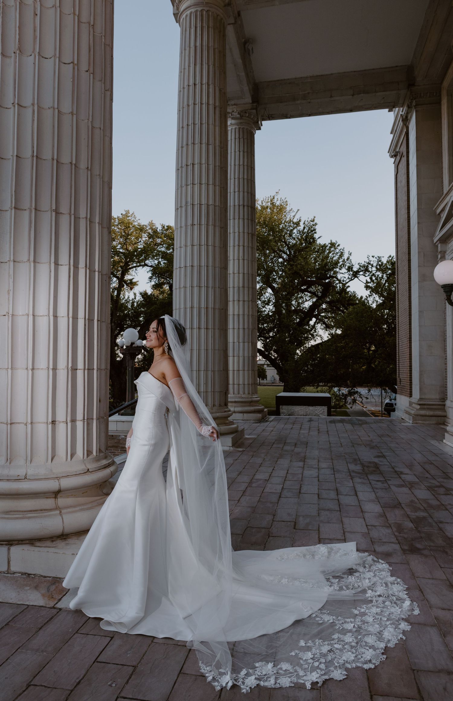 An elegant bridal portrait featuring a long lace-trimmed wedding dress among grand white columns of a historic building.