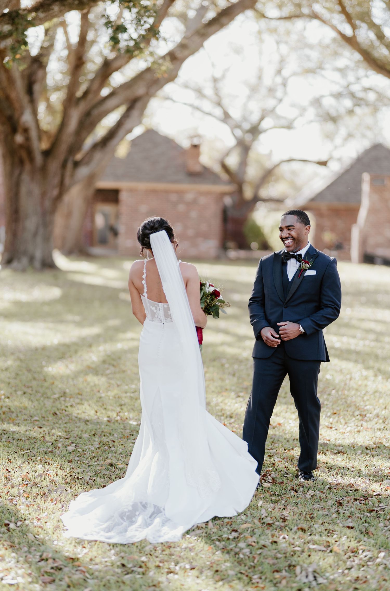 A romantic outdoor wedding scene with a bride in a white lace gown and groom in navy suit standing among large trees.