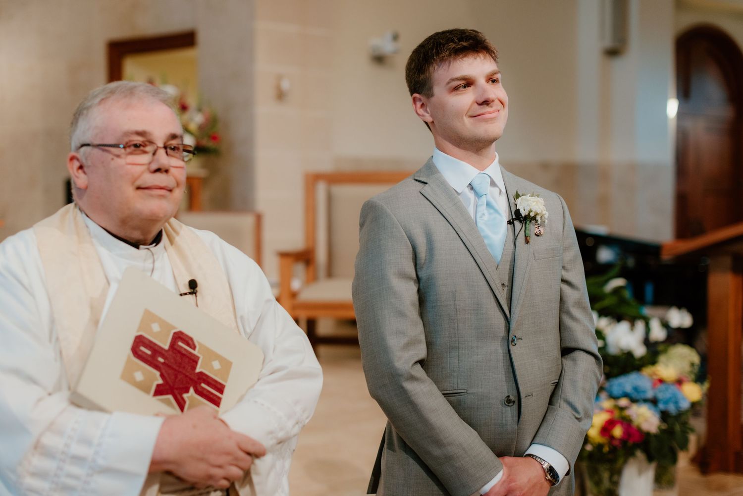 A priest in traditional vestments and a young man in a gray suit stand together during a church ceremony.