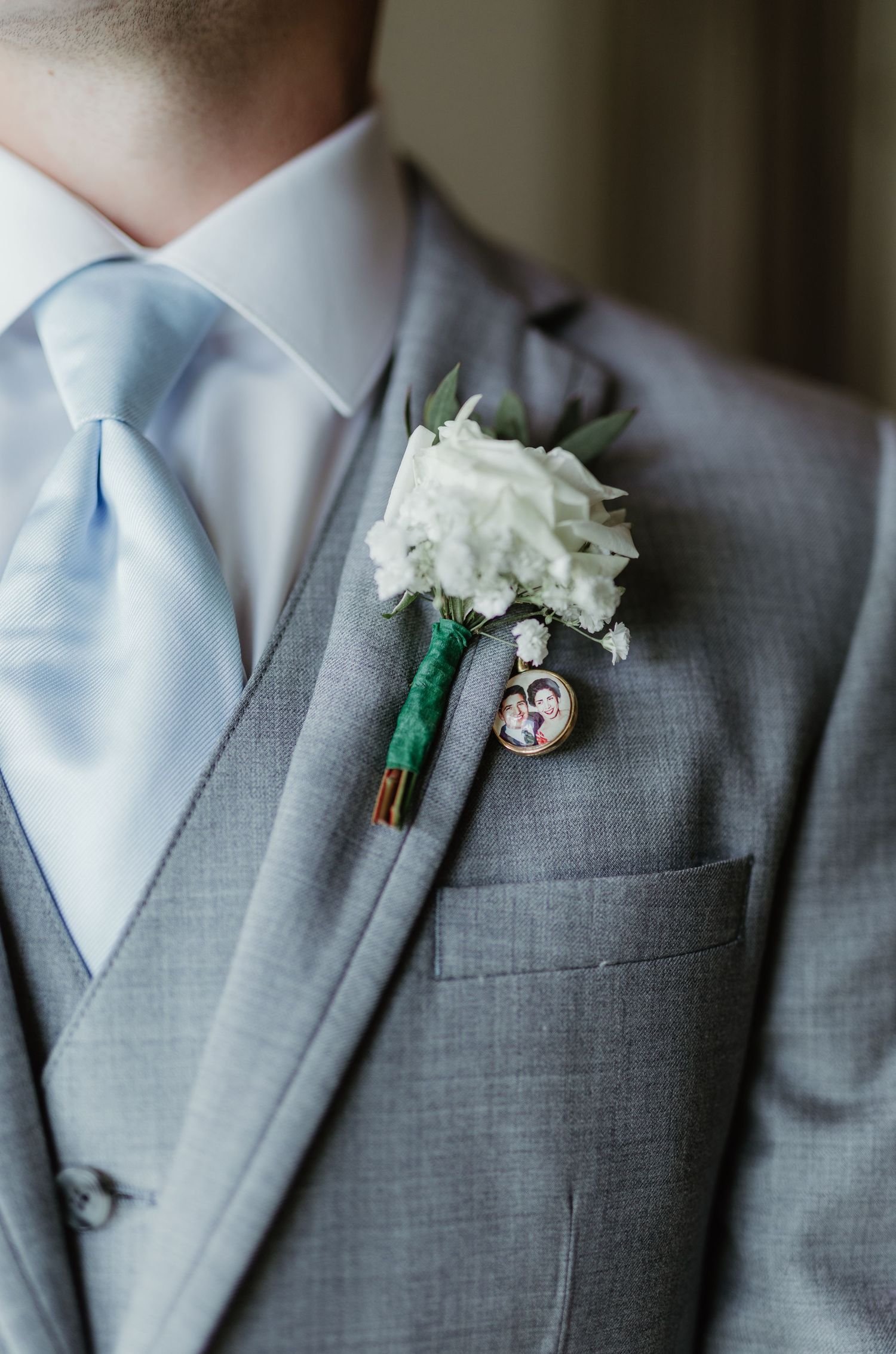 A white carnation boutonniere with a small photo charm pinned to a light gray suit jacket and baby blue tie.