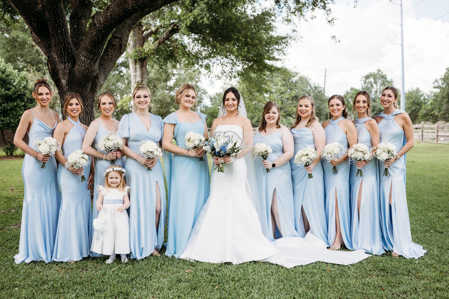 Wedding party poses outdoors in matching light blue flowing dresses with bouquets under a large shady tree.