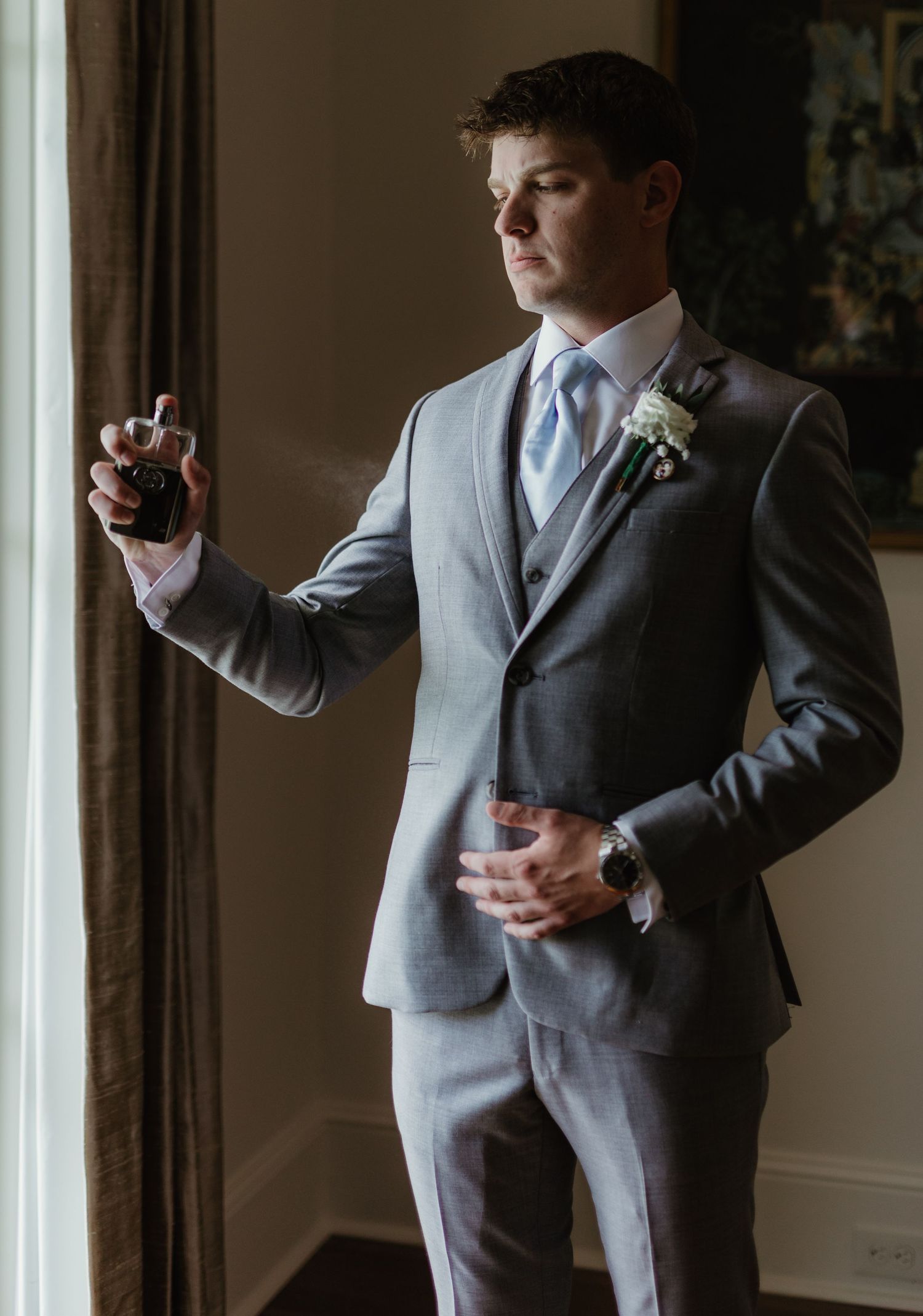 Groom in light grey suit and blue tie holds a drink while standing by window in moody lighting.