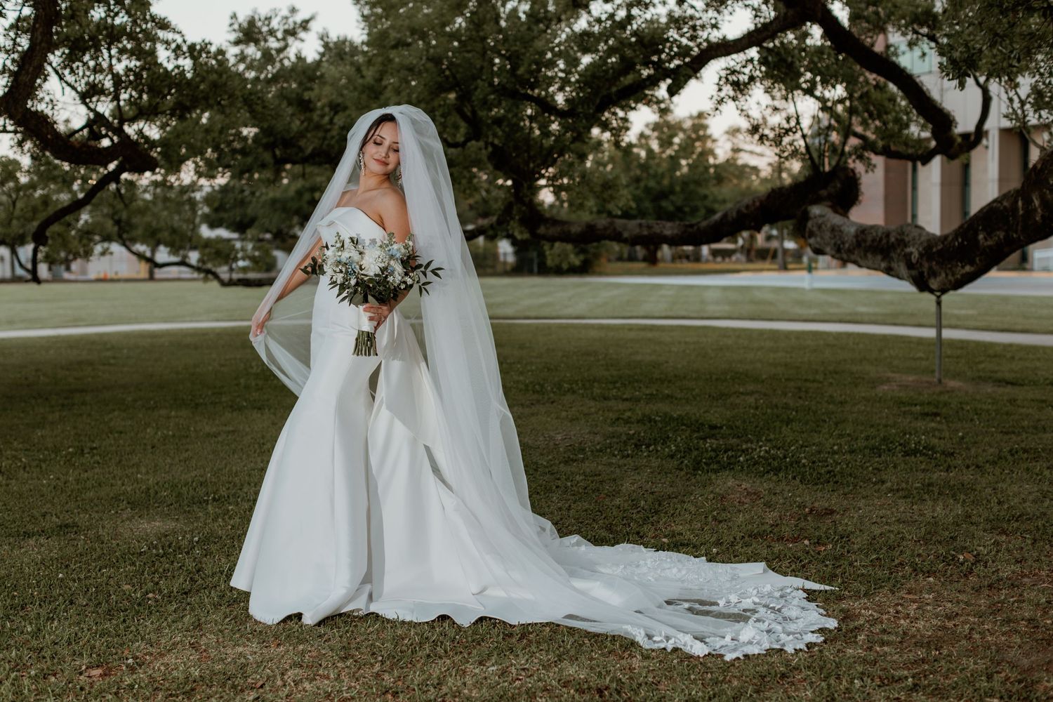 A stunning wedding gown with a flowing cathedral veil and white bouquet is captured on a grassy lawn surrounded by trees.