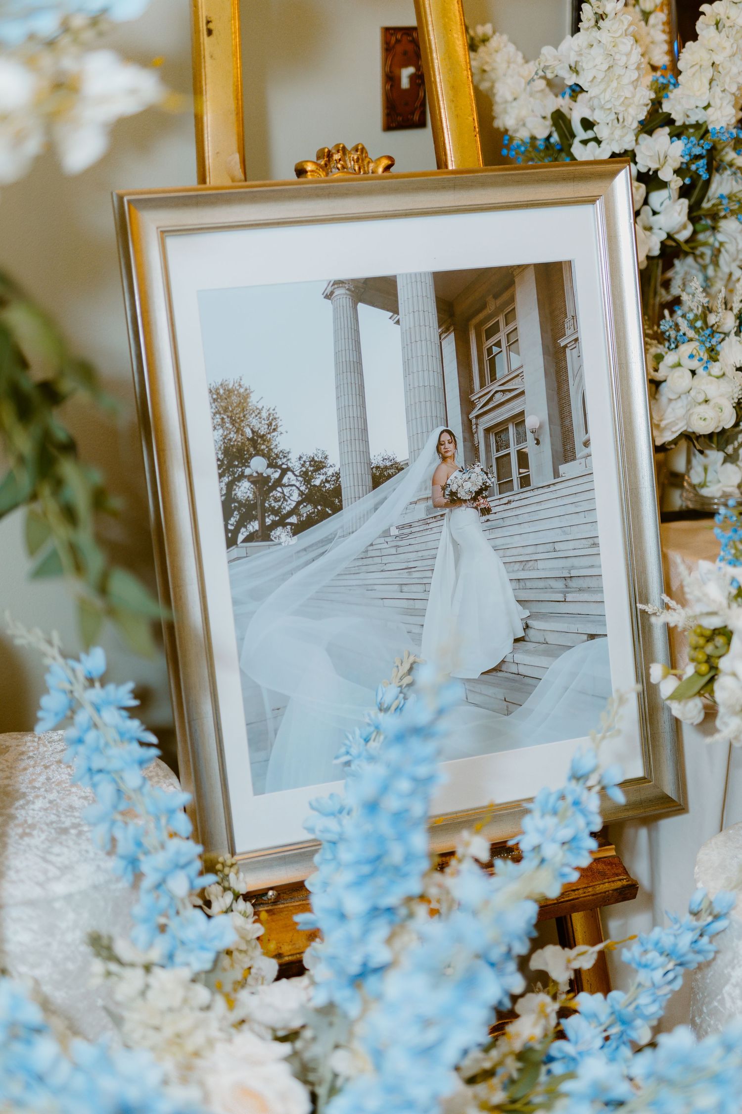 Framed wedding photo displayed on easel surrounded by blue and white delphinium and floral arrangements.