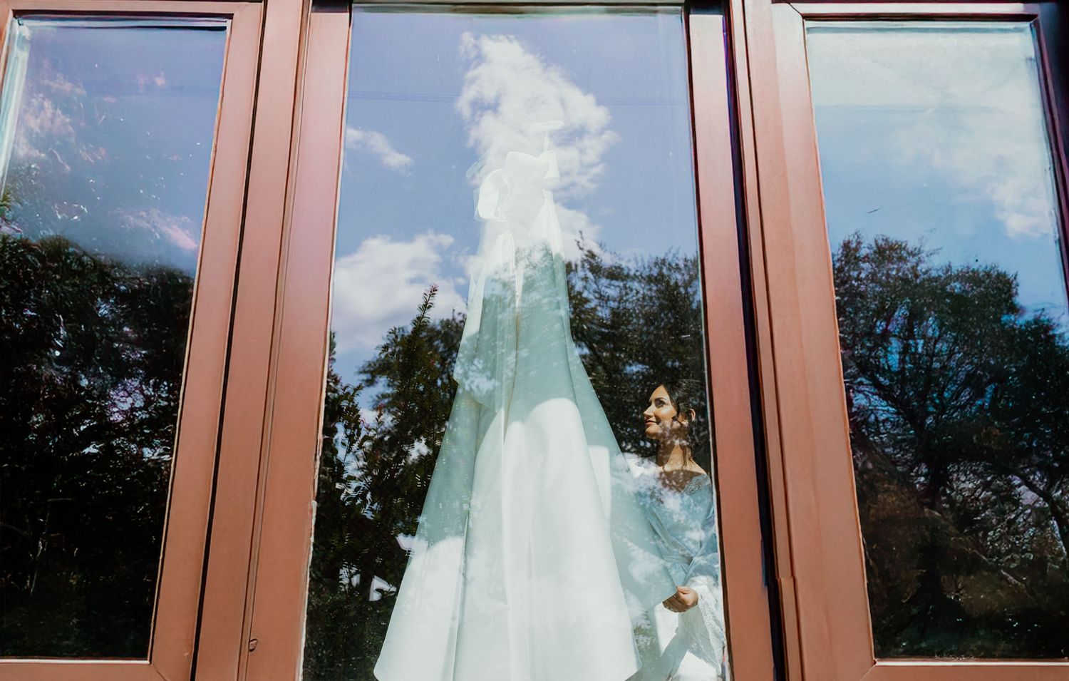 Wedding dress displayed in window reflection with copper-colored frame and trees visible in background.