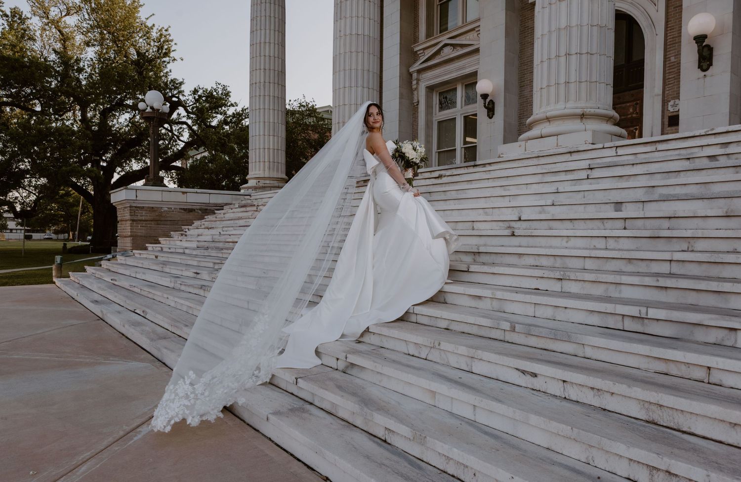 A bride in a flowing white gown and dramatic long veil descends elegant stone steps of a grand columned building.