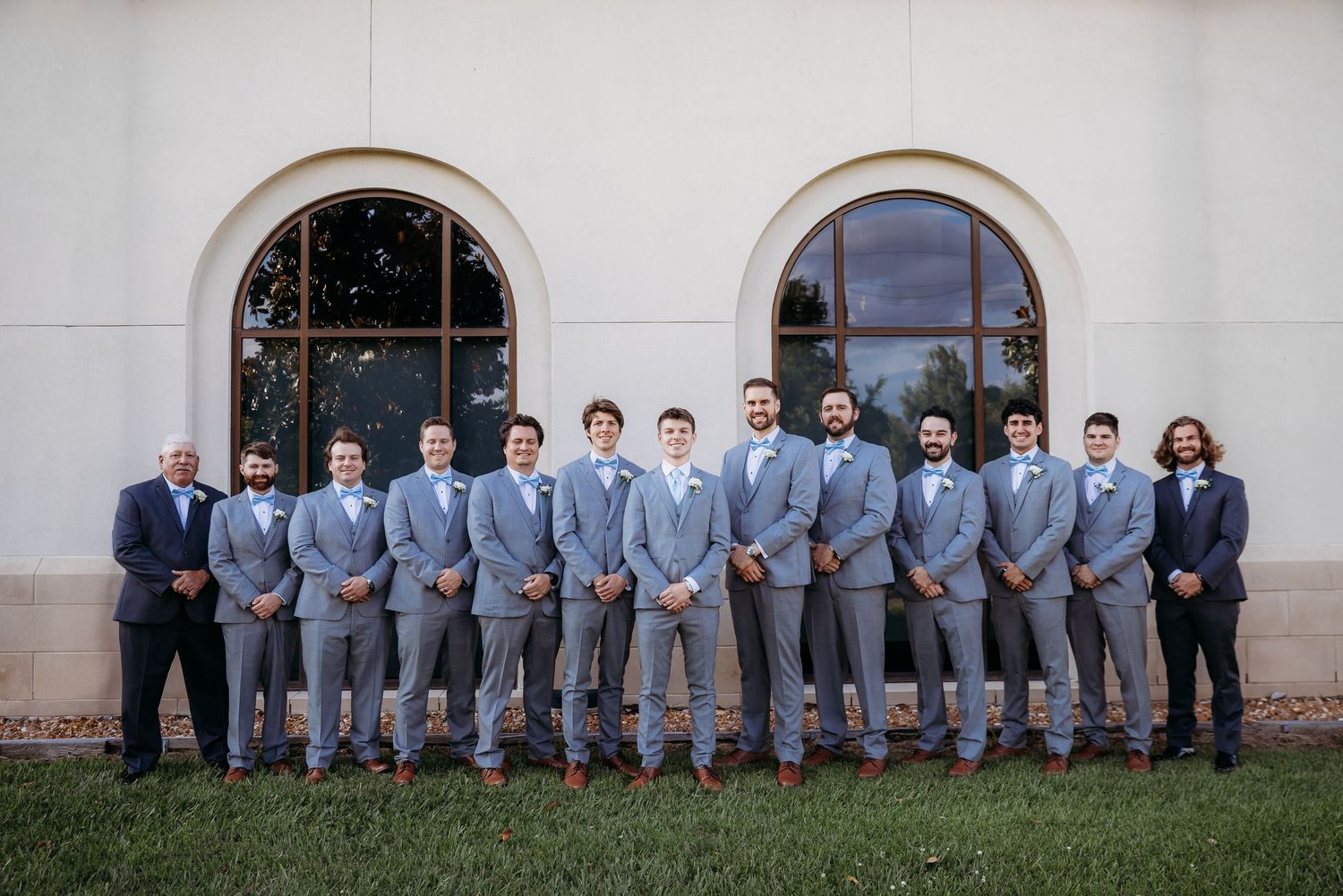 Wedding party of groomsmen wearing matching light gray suits lined up against white building with arched windows.