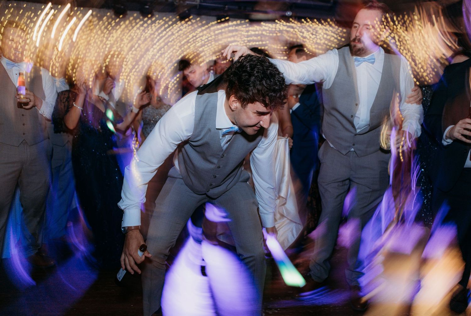 Wedding guests dancing energetically on a dance floor illuminated by colorful lights and motion blur effects.
