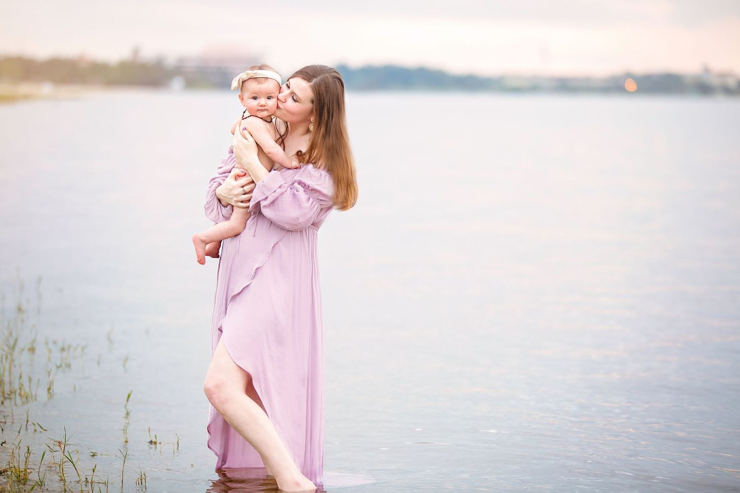 A family in pink outfits poses along a serene lakeside setting with tall grass and water views at sunset.