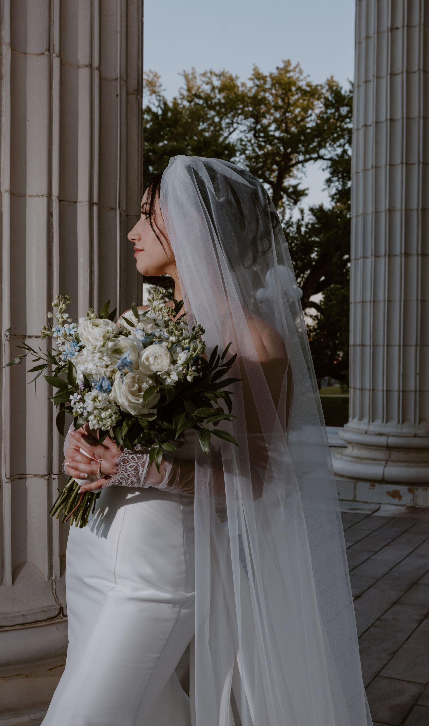 A bride in a sleek white dress and veil holds a lush bouquet of white flowers against classic stone columns.