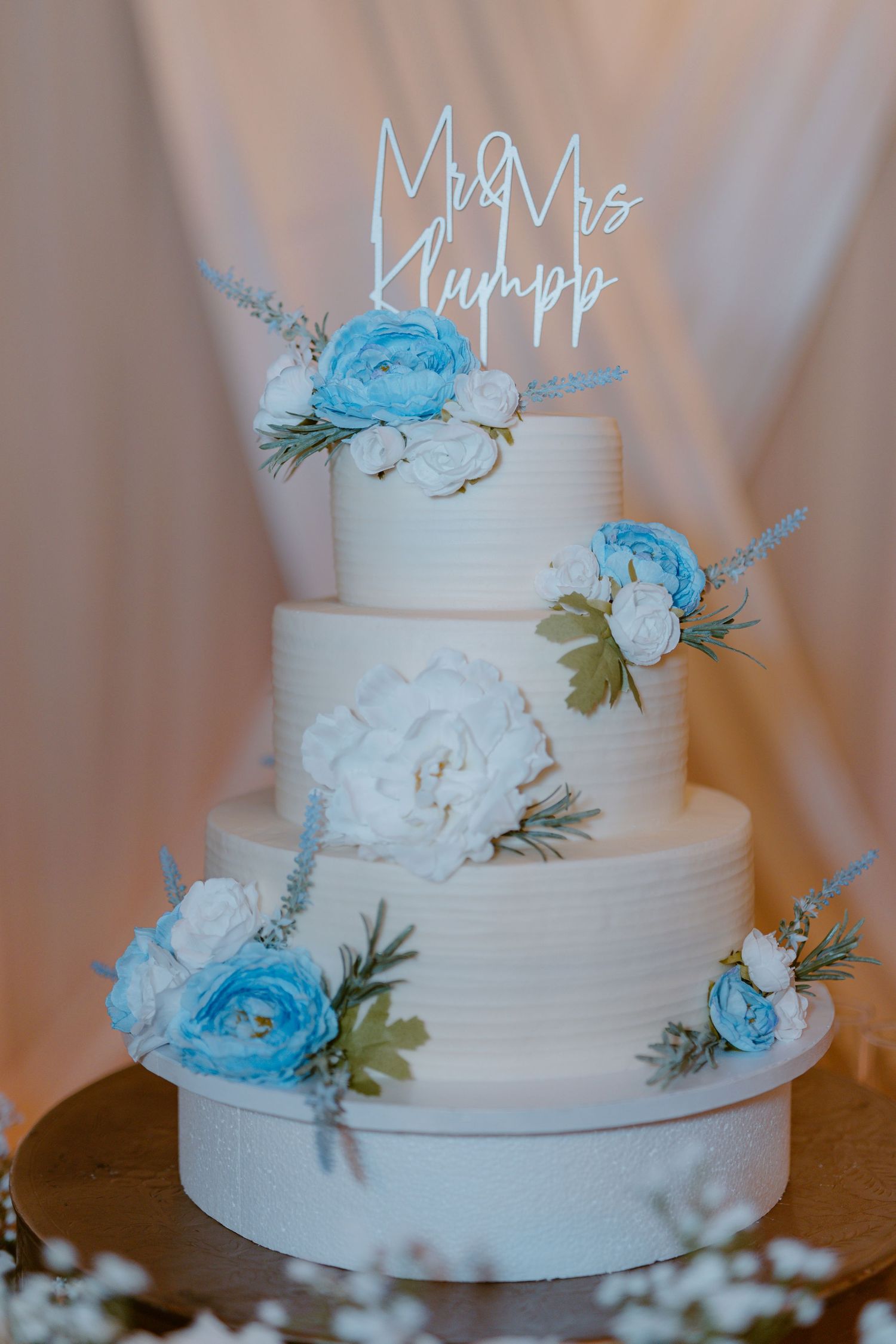 An elegant white wedding cake decorated with light blue roses and greenery, topped with a 'Mr & Mrs Khumpp' sign.