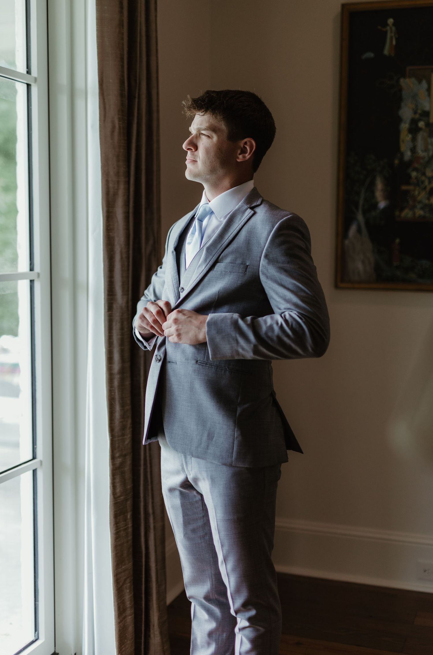 A person in a gray suit stands contemplatively by a window with natural light streaming in, creating a moody atmosphere.