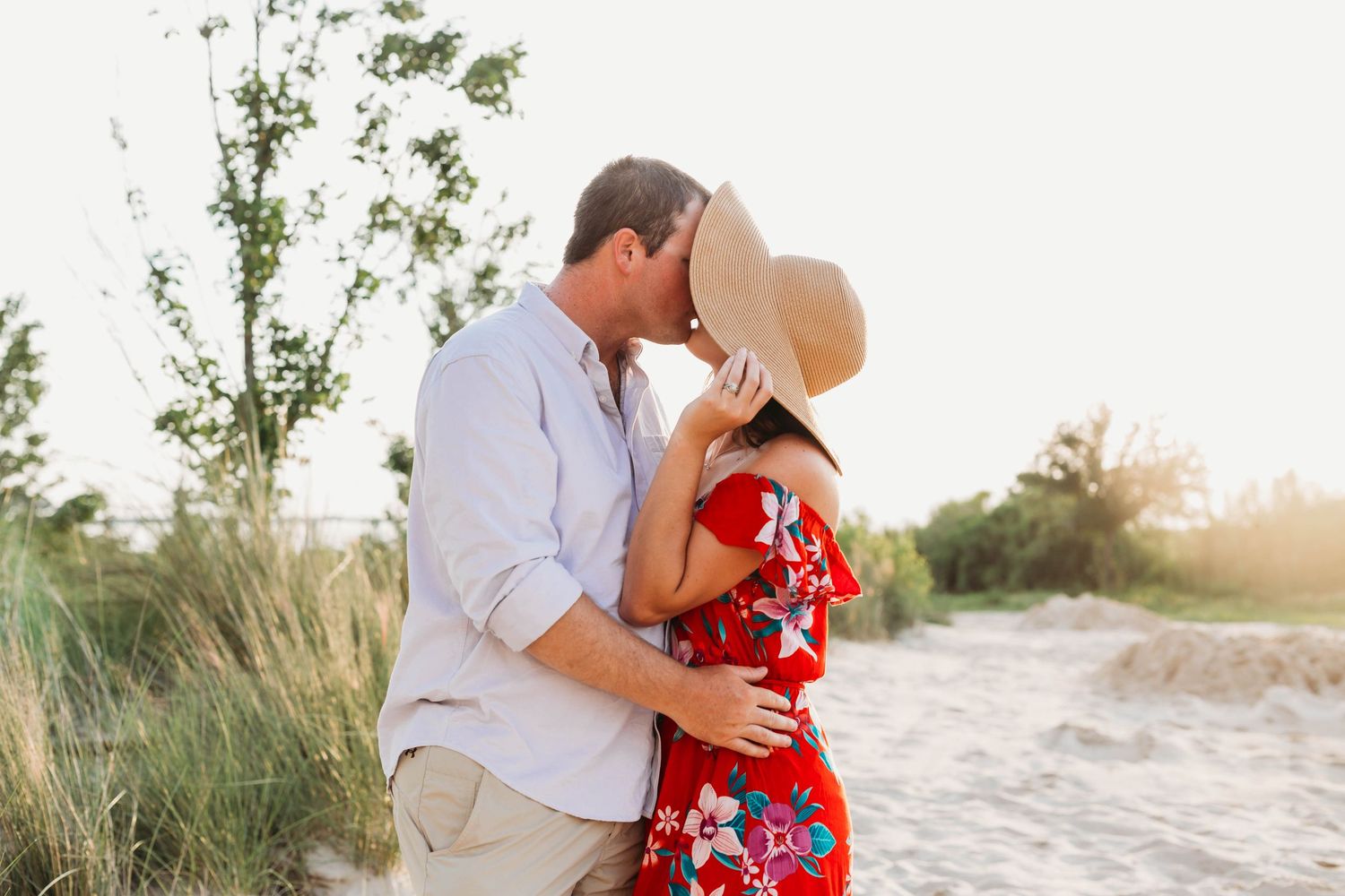 Romantic couple embraces on sandy beach path at sunset with tall grass swaying in background.