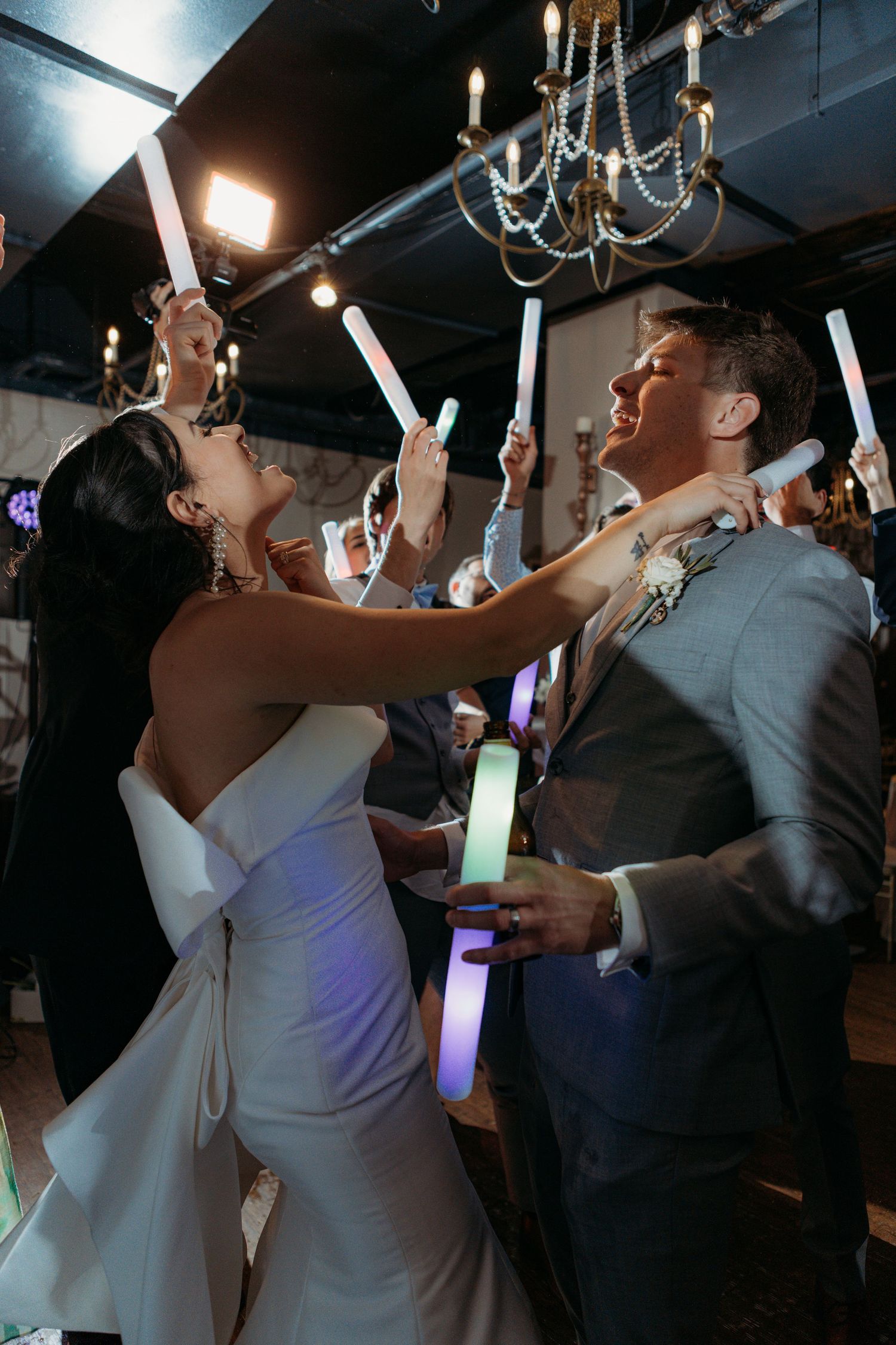 Wedding couple holds up glowing LED foam sticks while dancing under a chandelier at their reception celebration.