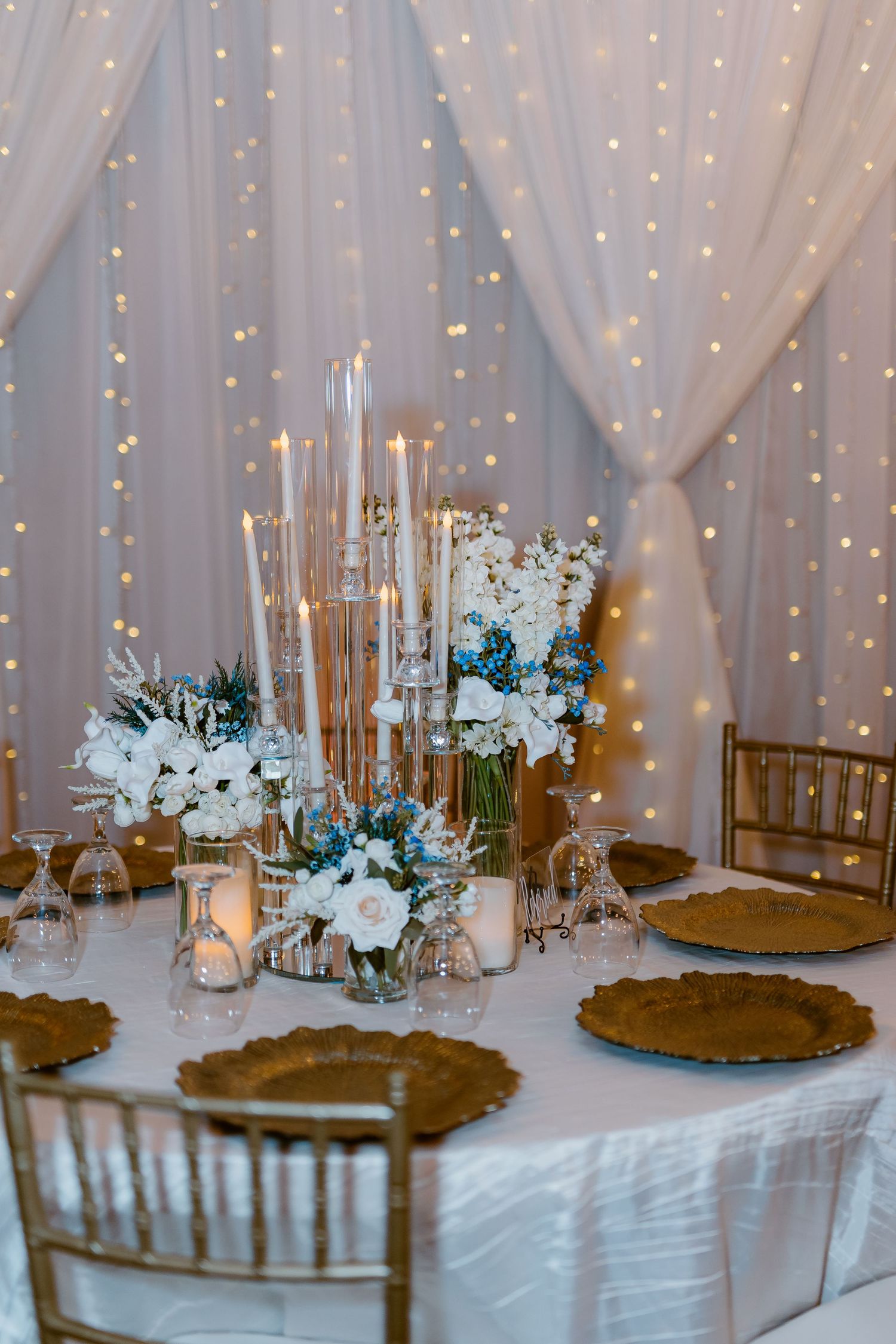 Elegant wedding table setup with tall gold candles, white flowers and draped fabric backdrop with twinkling fairy lights.