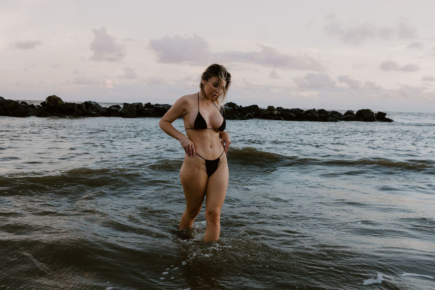 A person stands in shallow ocean water at dusk with a rocky breakwater visible in the background.