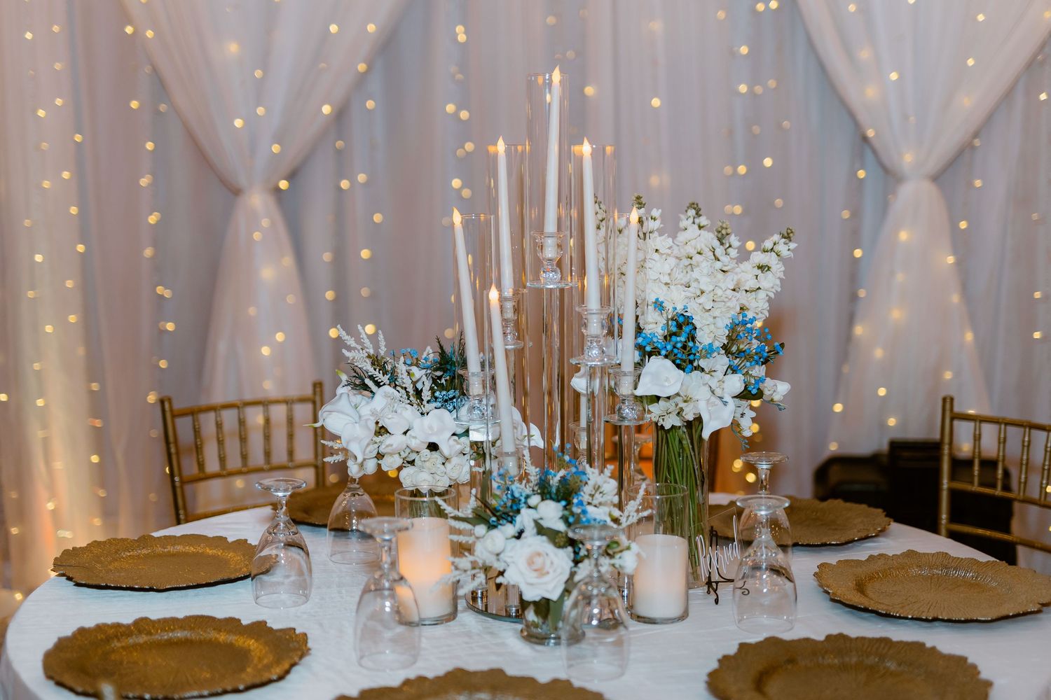 Elegant wedding reception table setting with teal flowers, tall candles, and gold charger plates against a twinkling backdrop.