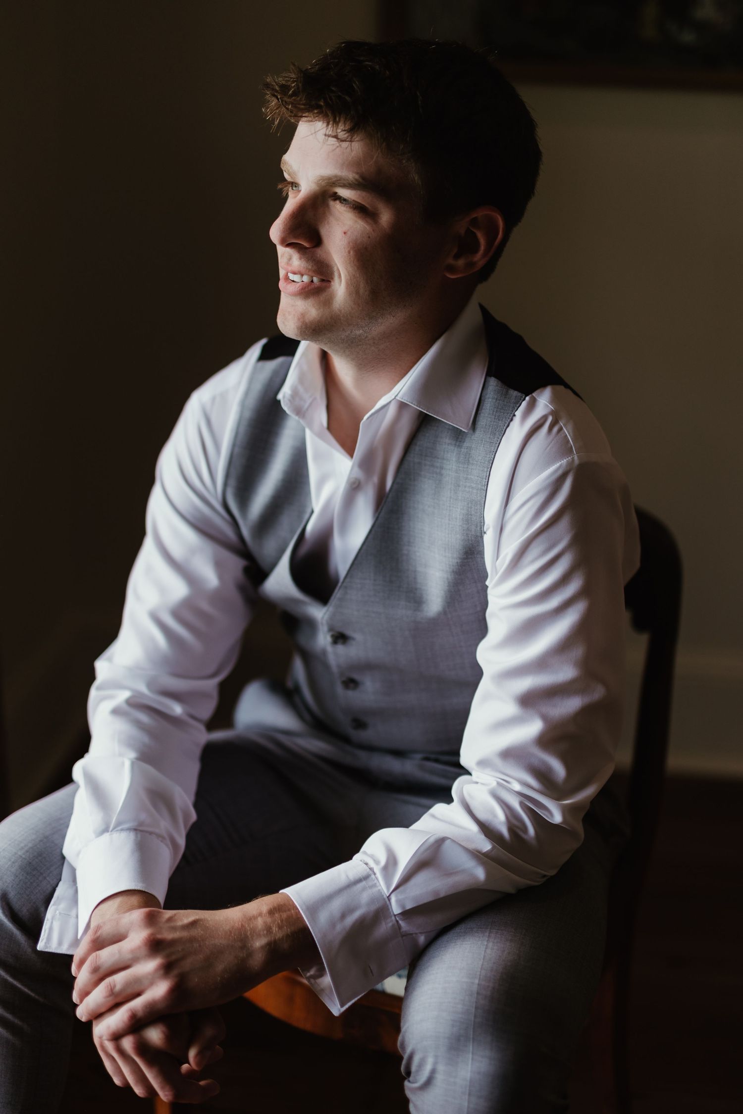 A groom in a white shirt and grey vest sits looking to the side in contemplative natural lighting.