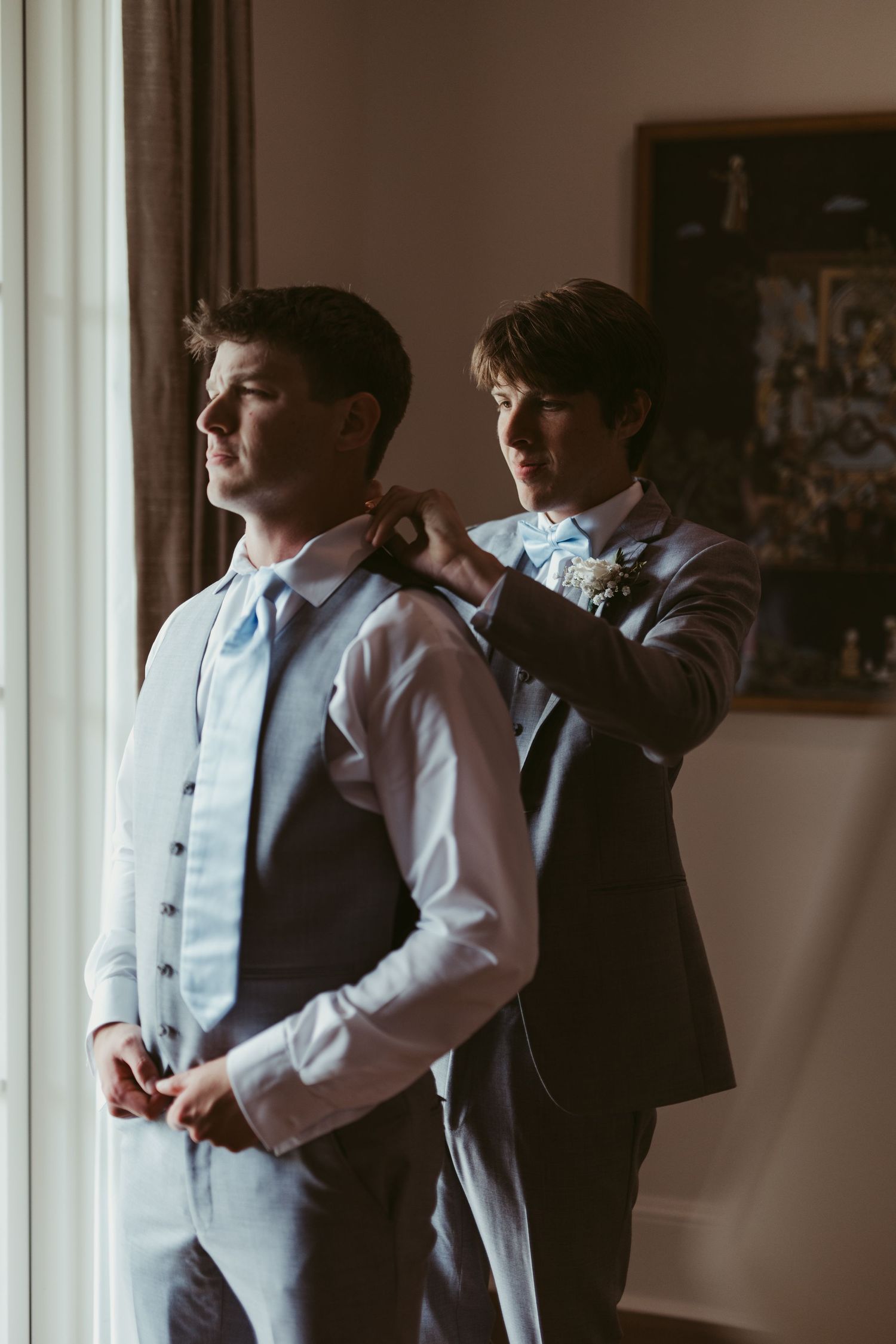 Two groomsmen in formal attire help each other get dressed by a window during wedding preparations.