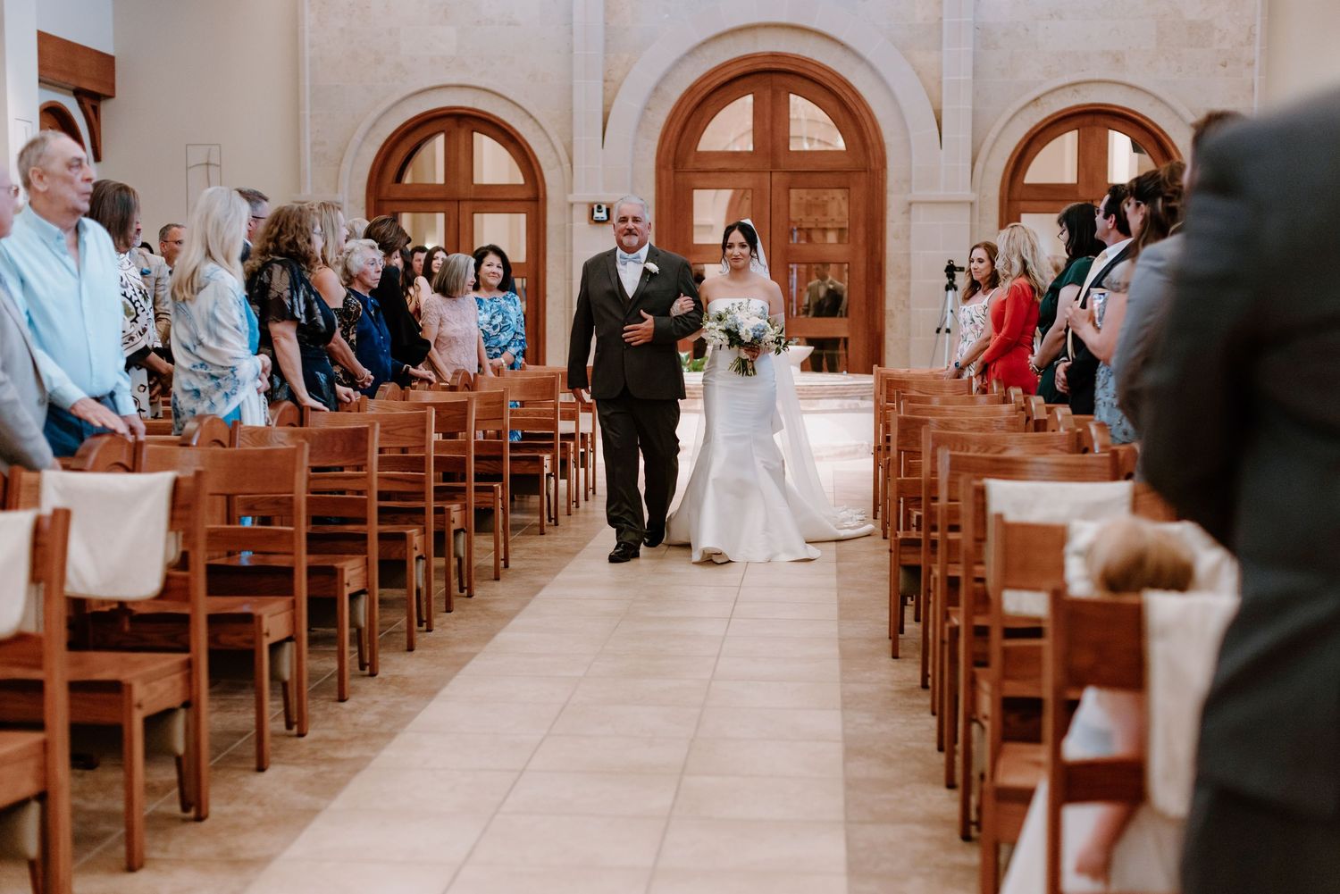 A bride walks down a church aisle with wooden pews lined with wedding guests on either side.