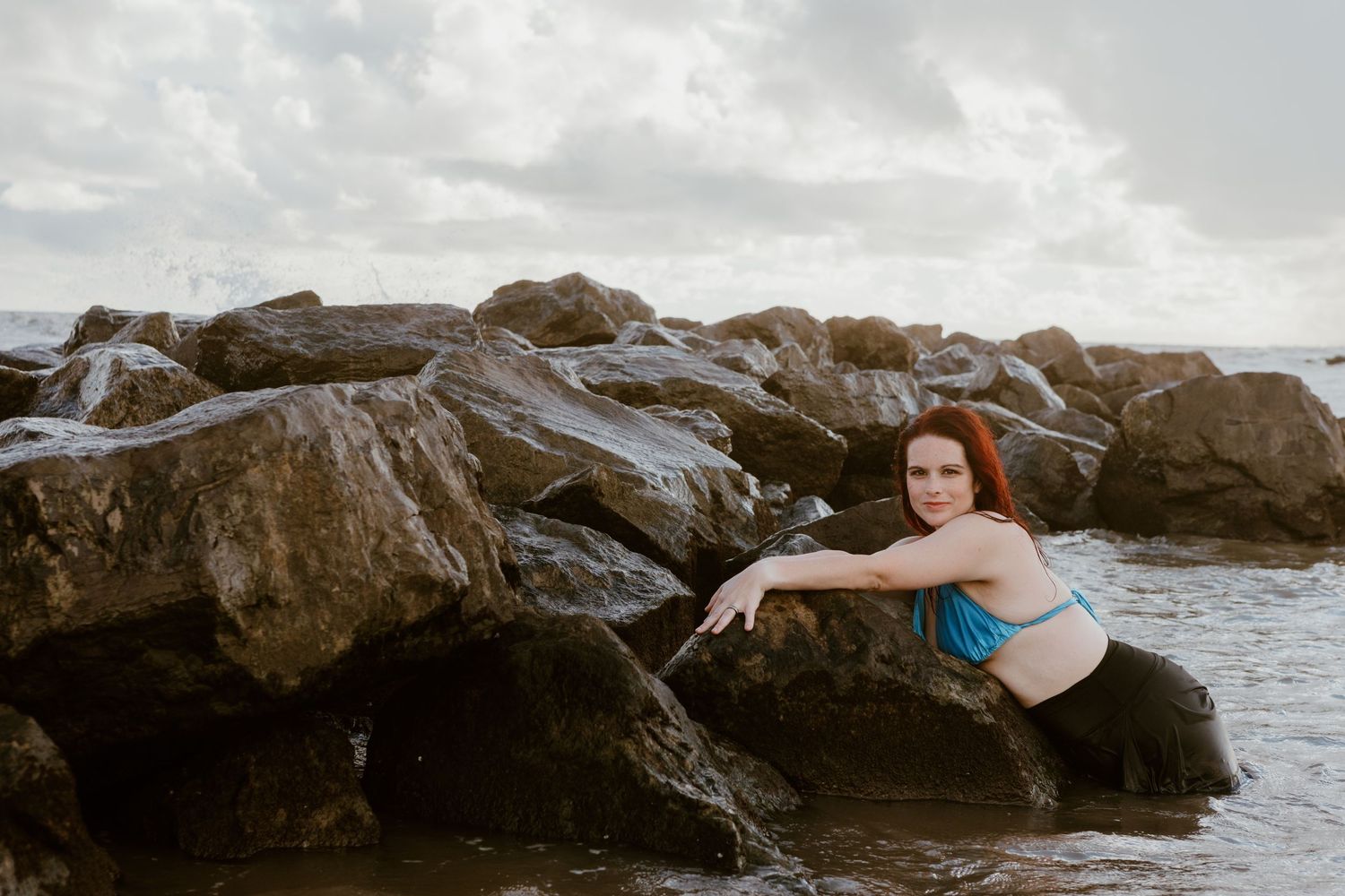 A person in a blue swimsuit lounges on seaside rocks with waves crashing around them along a rocky coast.