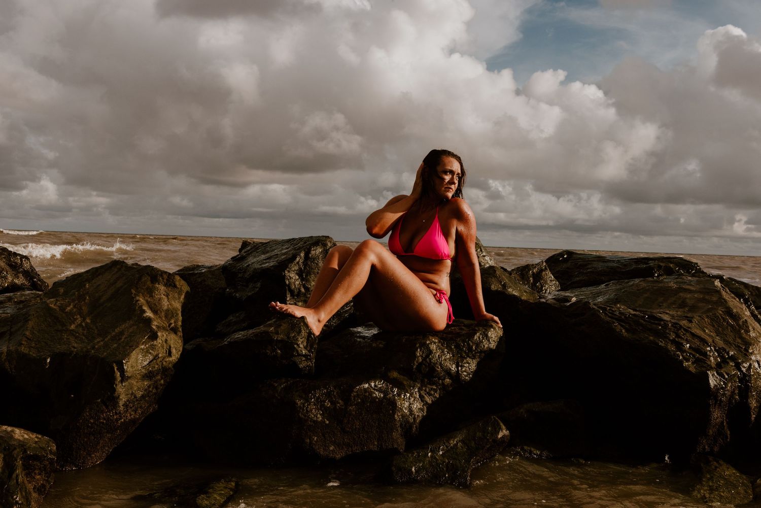A figure in a pink swimsuit sits atop dramatic black volcanic rocks against a cloudy sky at sunset.
