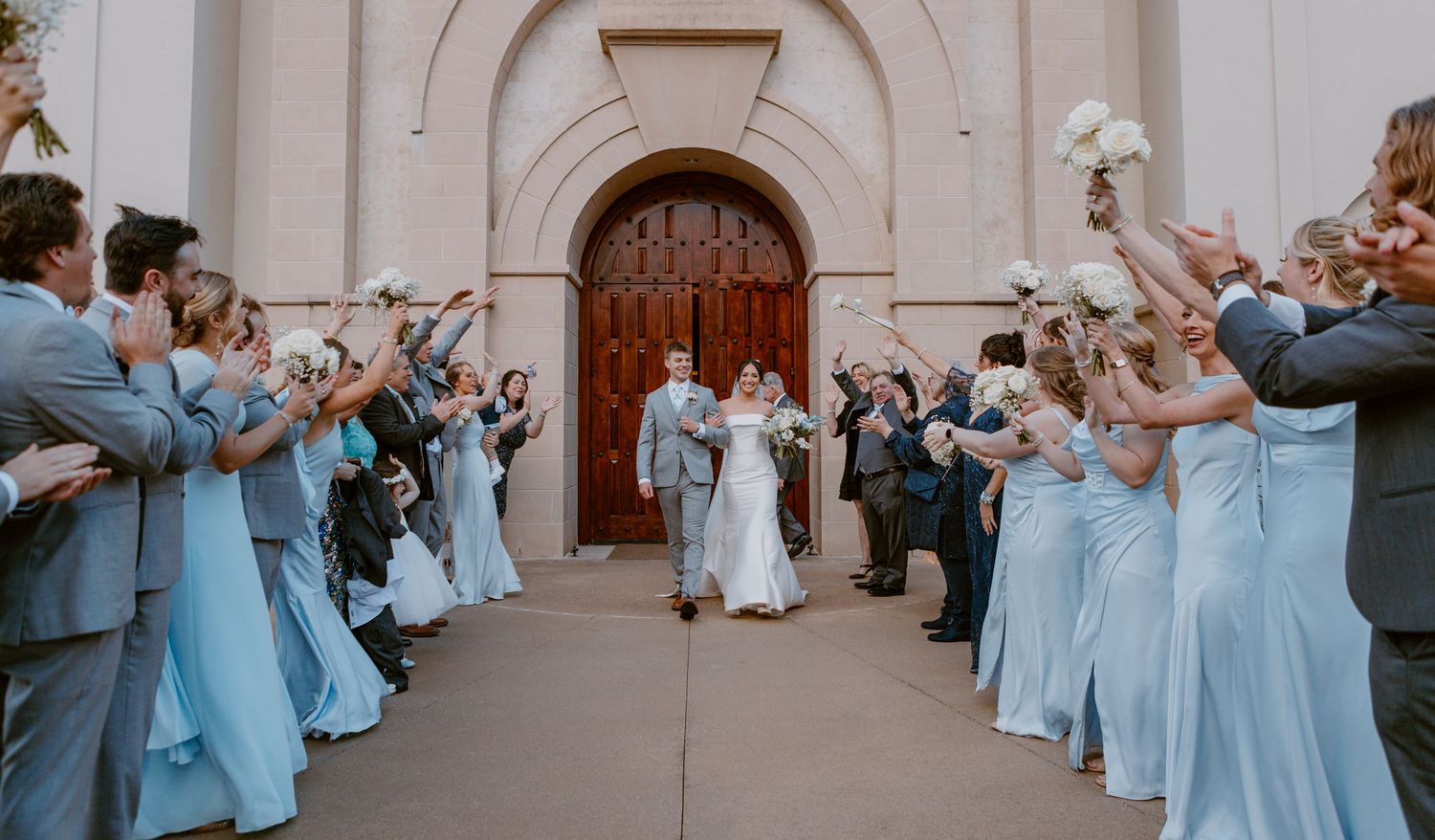 Wedding party in powder blue dresses and gray suits creates aisle for couple exiting church ceremony through arched doorway.