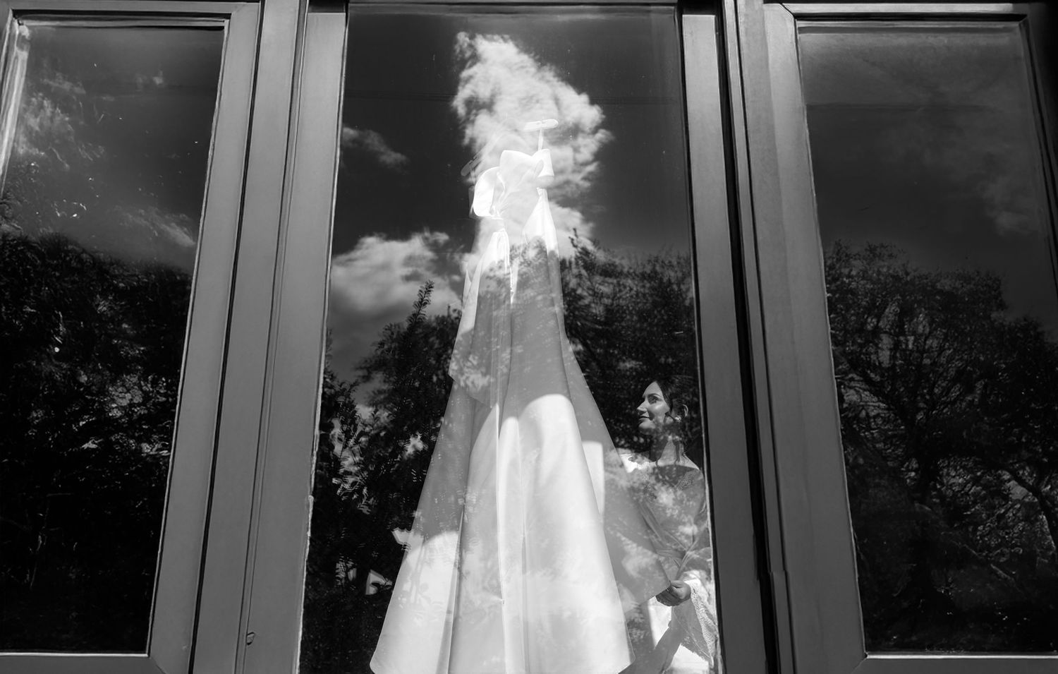A black and white photo of a wedding dress displayed in a window with dramatic reflections and shadows.