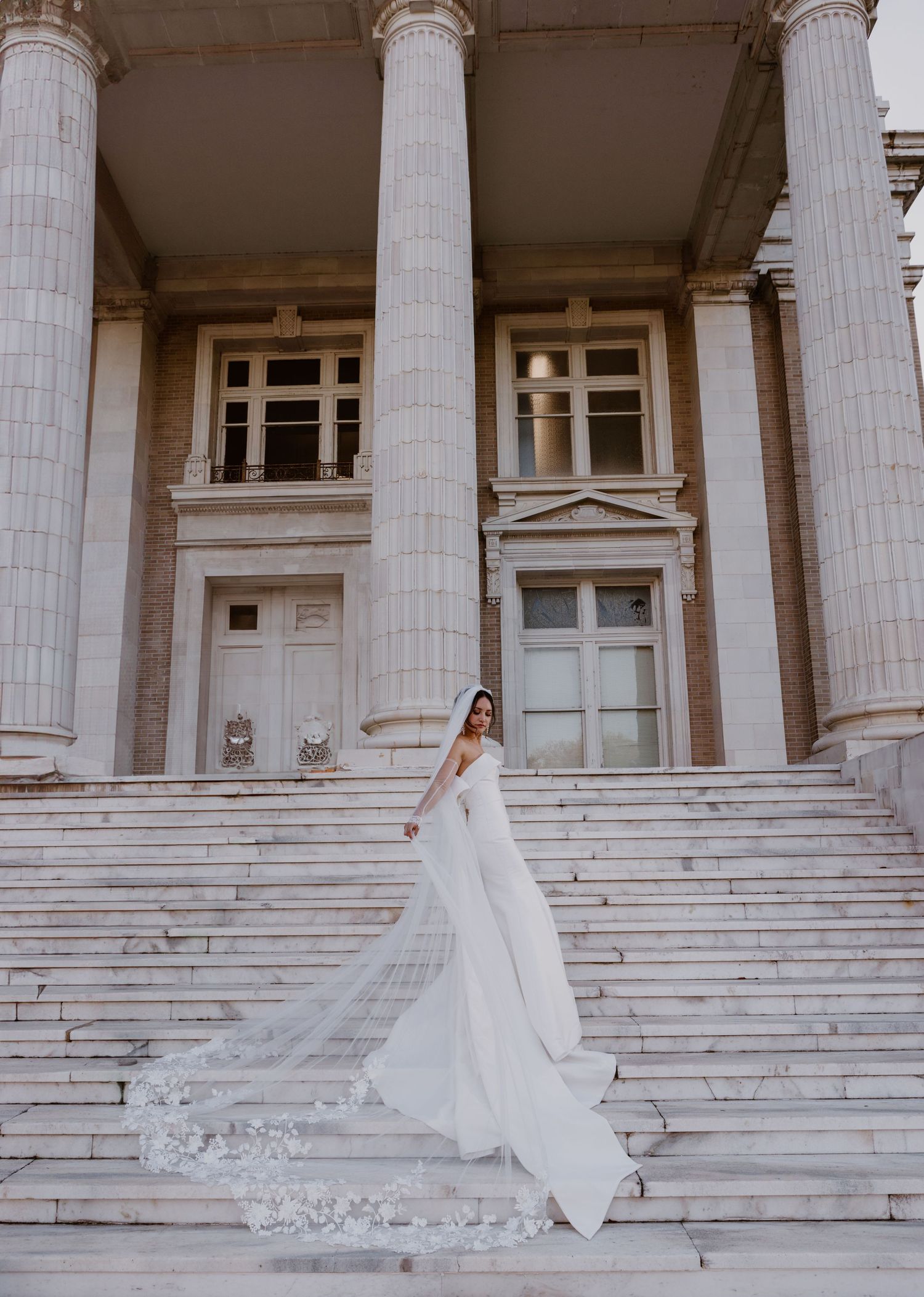 A flowing white wedding gown with long train cascades down marble steps of a grand building with classical columns.
