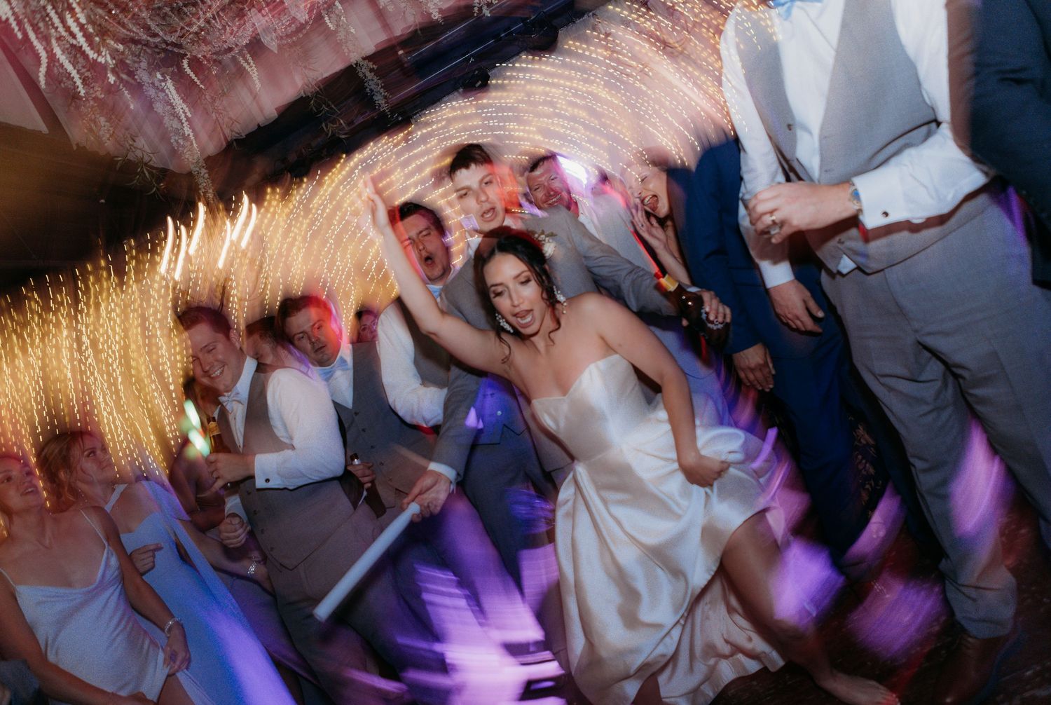 Wedding reception dance party with colorful purple lighting and energetic dancing.