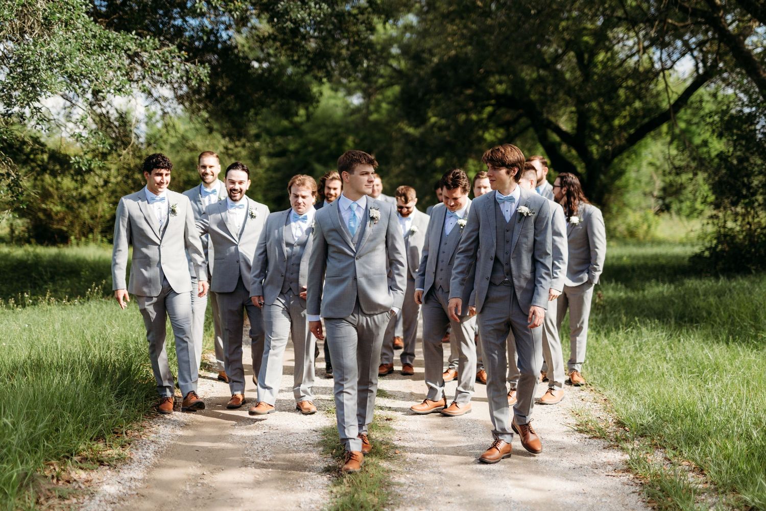 A group in coordinated grey suits walking together down a path in a sunlit outdoor setting.
