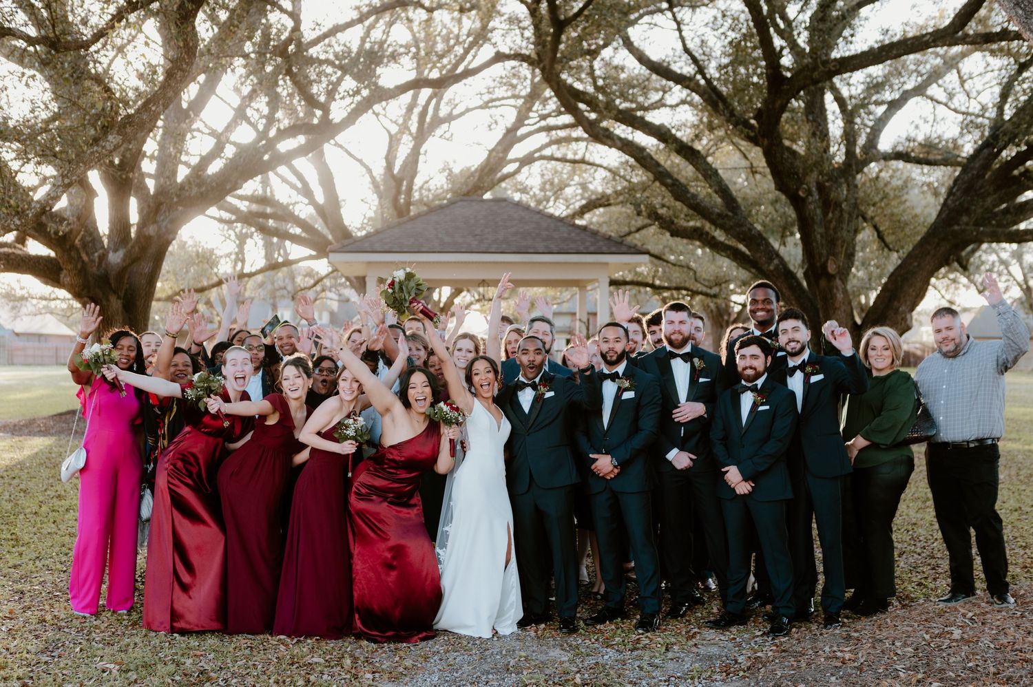 Large wedding party poses together outdoors with bridesmaids in burgundy dresses and groomsmen in black suits at sunset.