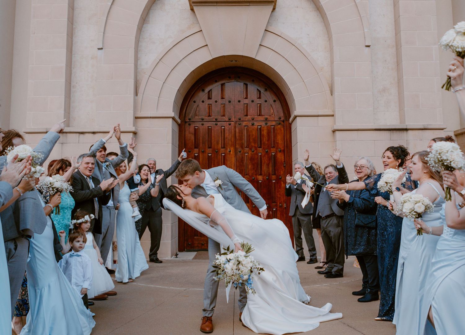 Wedding party in light blue dresses celebrates as newlyweds share dramatic romantic dip kiss outside church entrance.