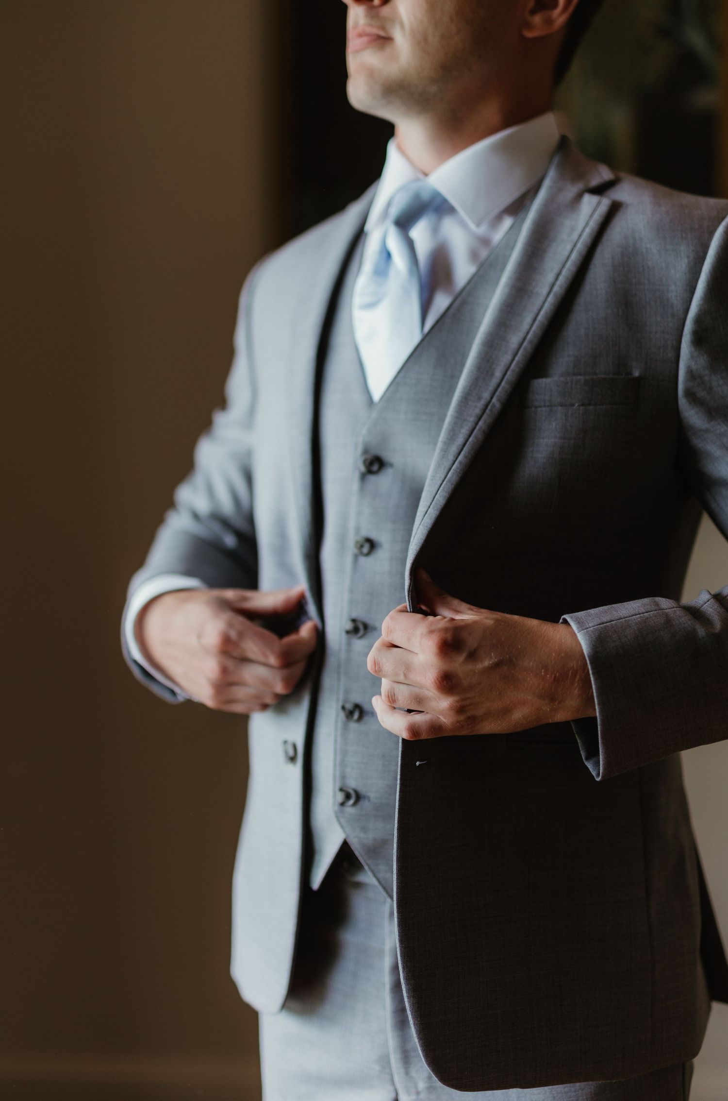 Close-up of groom adjusting his gray three-piece suit and light blue tie while getting ready.