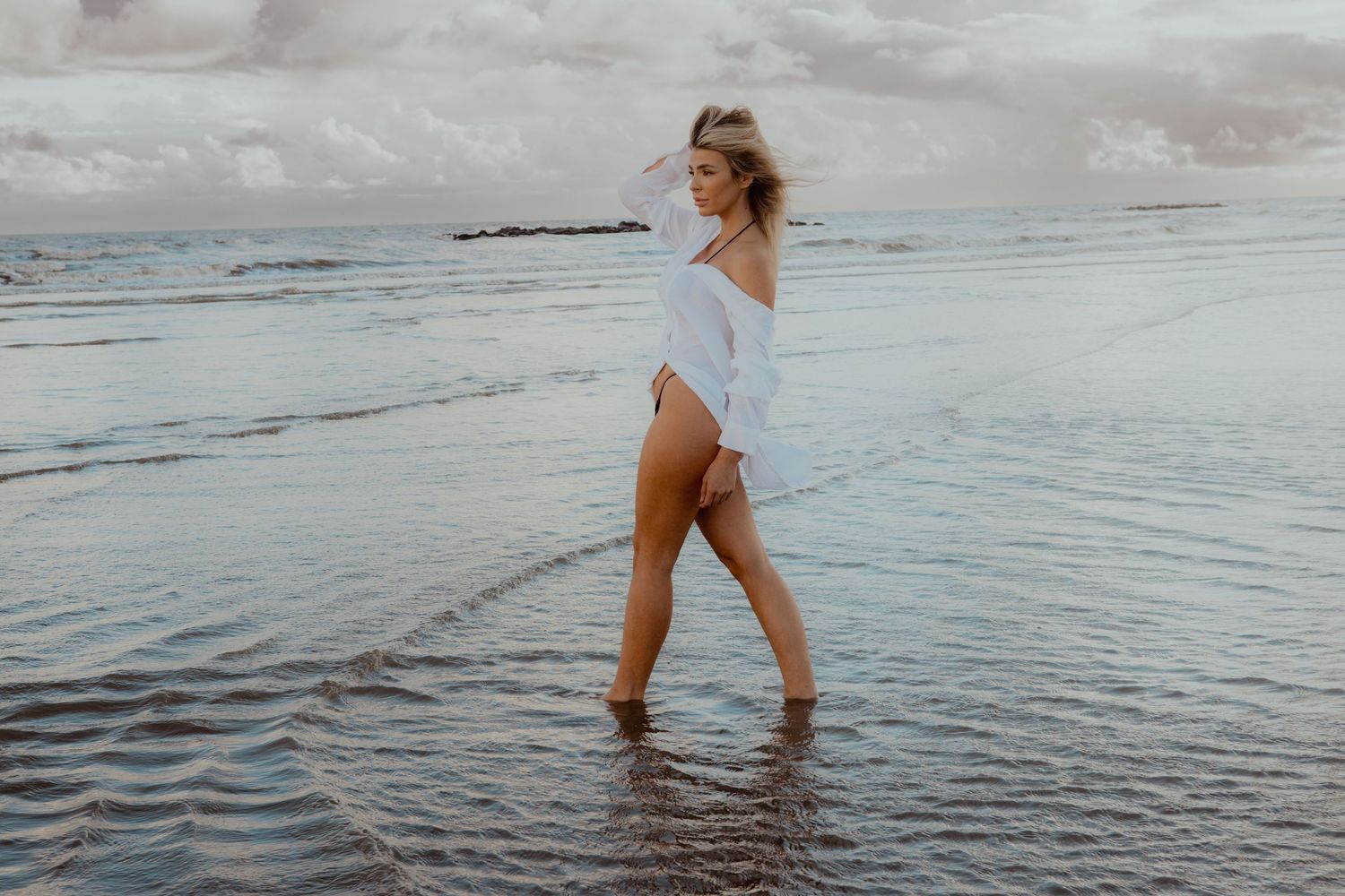 A person in a white shirt walks through shallow ocean water at sunset on a cloudy beach.
