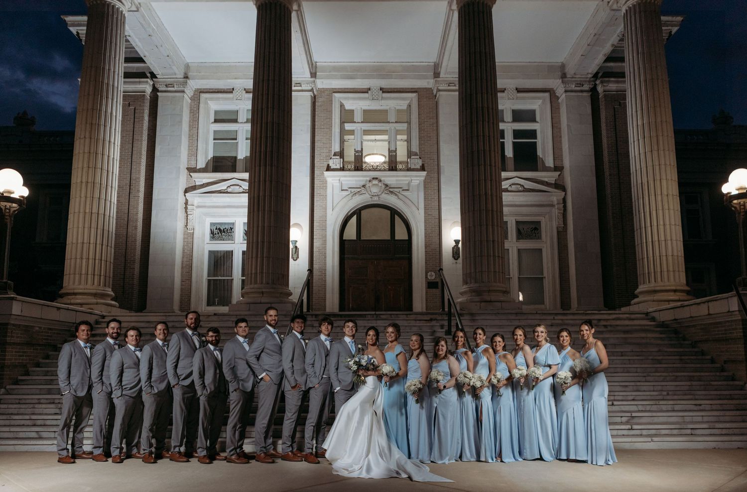 Wedding party in gray suits and light blue dresses pose on grand stone steps of elegant building at night.